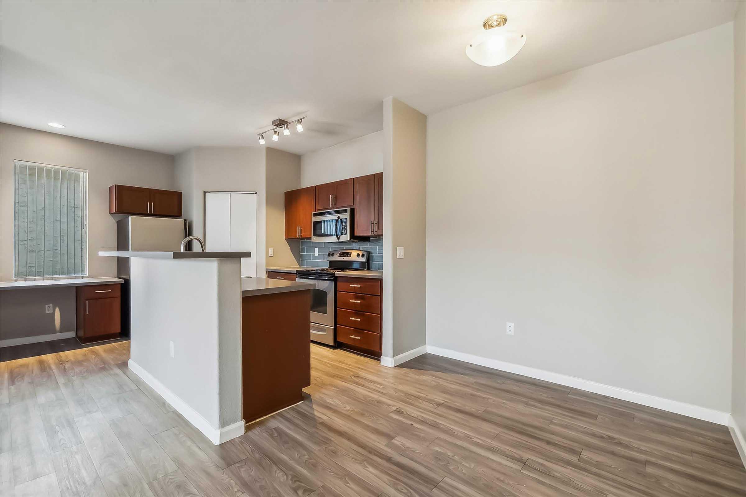 Modern kitchen and living area in an apartment. The kitchen features stainless steel appliances, brown wooden cabinets, and a light countertop. The flooring is light wood, and the walls are painted in neutral tones, creating a spacious and inviting atmosphere.