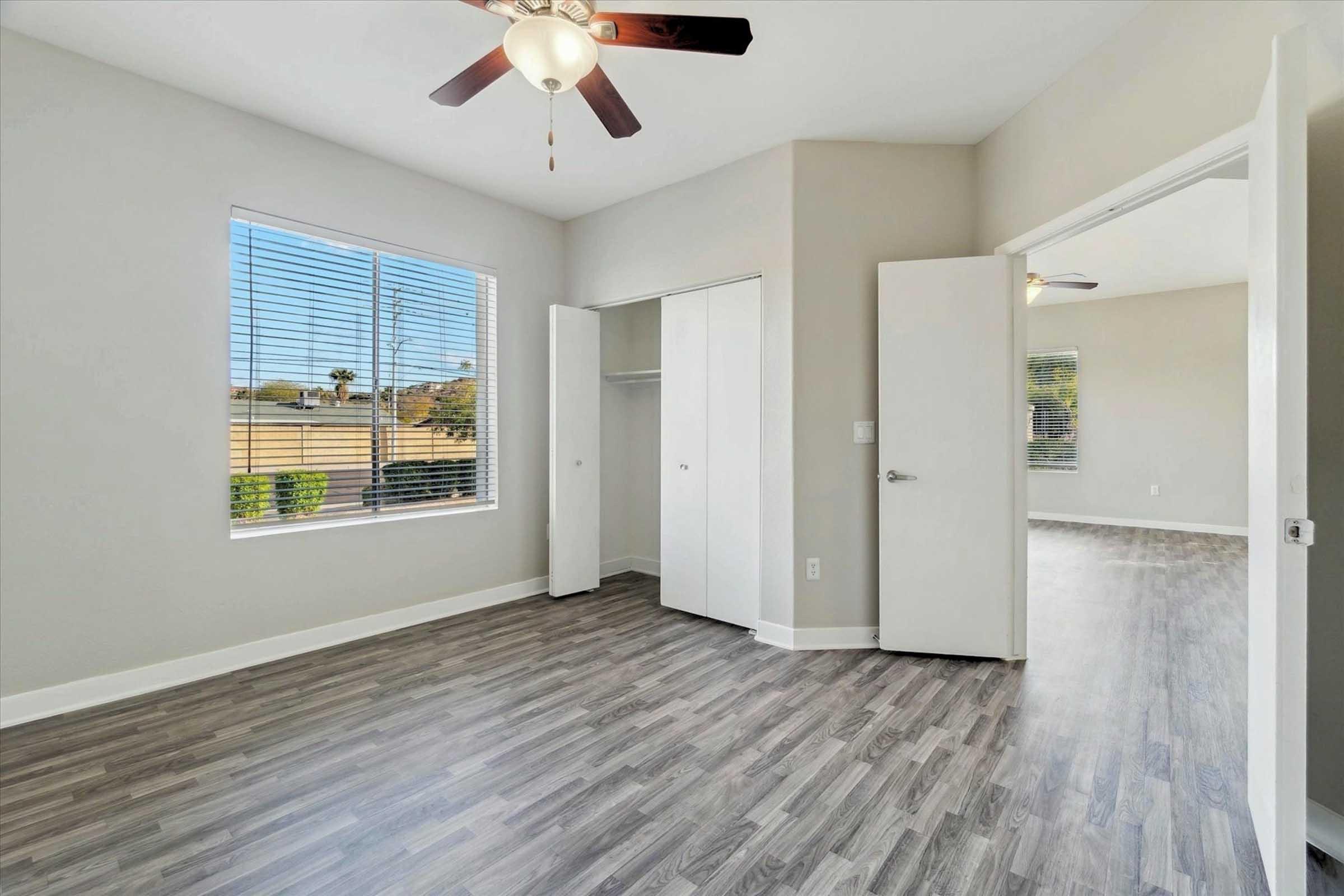 Bright, empty room featuring light gray walls, a ceiling fan, and vinyl flooring. Two white doors lead to a closet and another room, with large windows allowing natural light to flood in. The view includes greenery outside, creating a welcoming and spacious atmosphere.