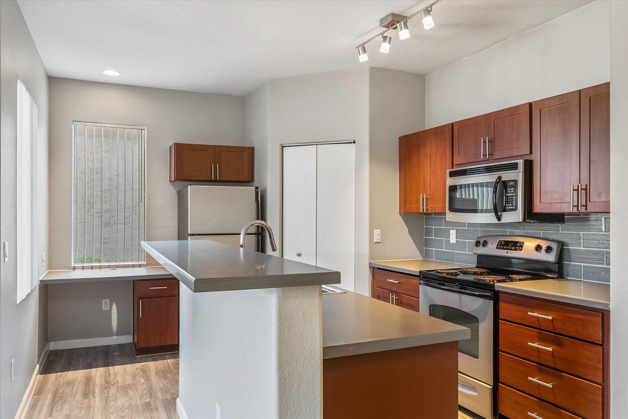 Modern kitchen featuring wooden cabinets, stainless steel appliances, and gray countertops. The layout includes a kitchen island with a sink, a stove, and a refrigerator. Natural light enters through a window, and the walls are painted in neutral tones.