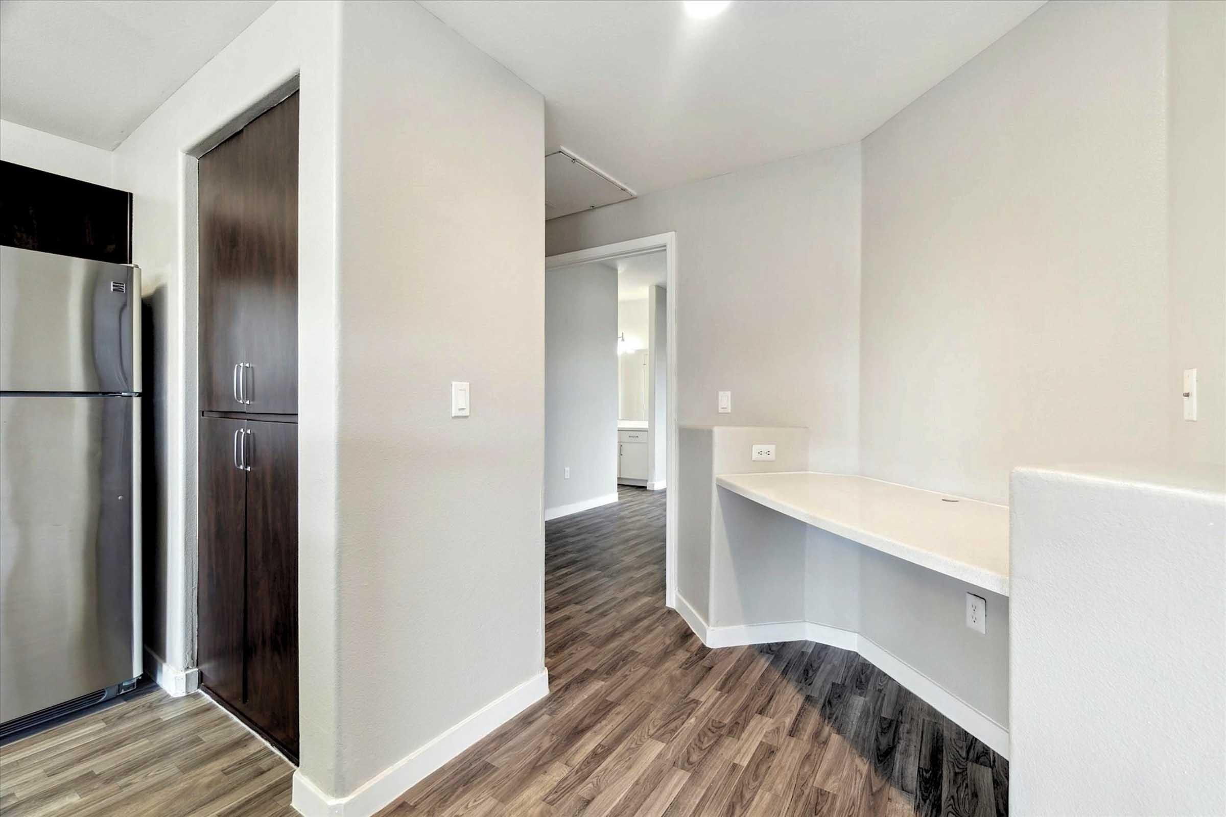 Bright and modern interior view of a kitchen area featuring stainless steel appliances, a dark wood pantry, and a small desk space against a white wall. The flooring is a trendy wood laminate, creating a warm and inviting atmosphere. The layout leads to an adjoining room with soft lighting.