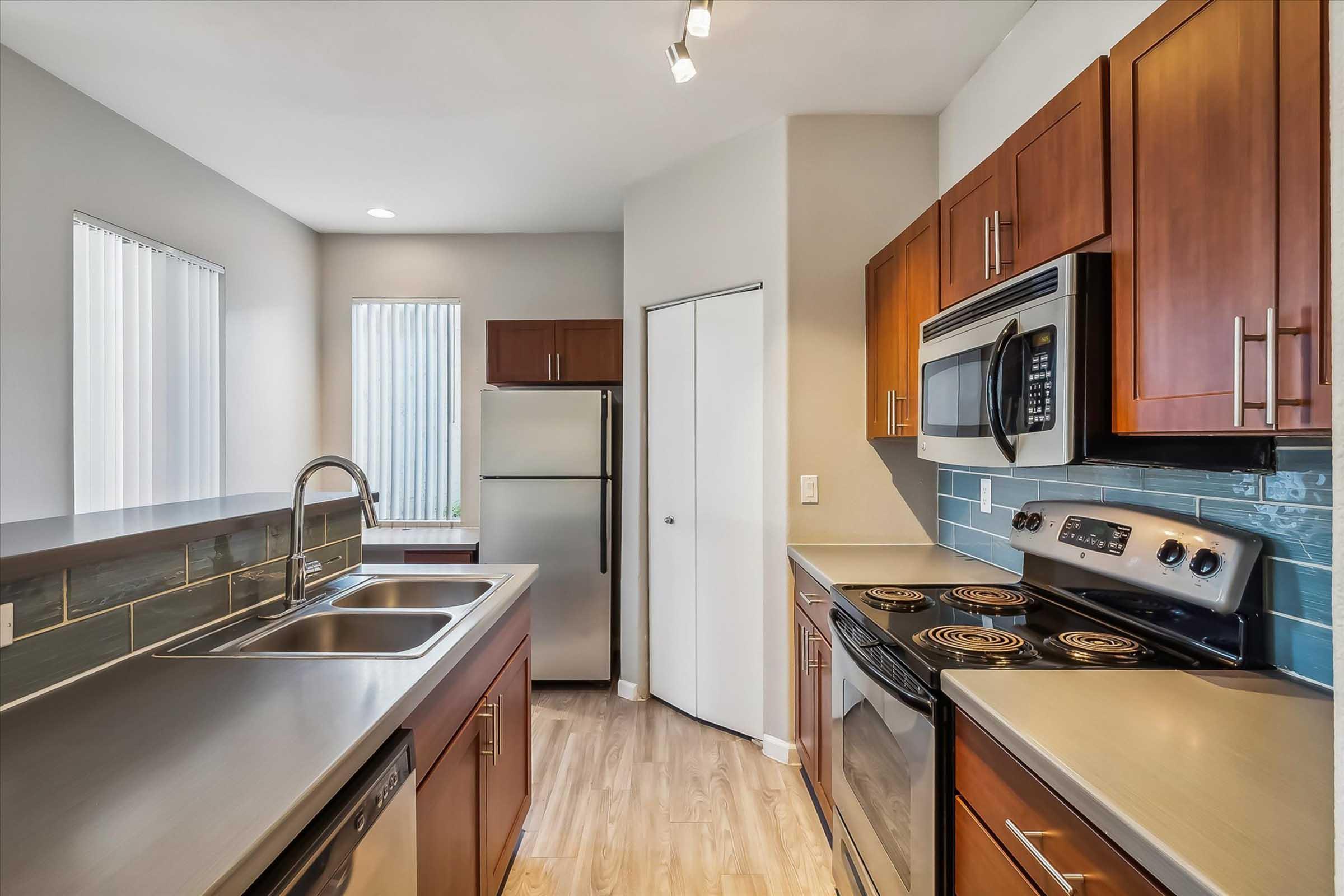 Modern kitchen featuring stainless steel appliances, including a microwave and oven. Dark wood cabinetry contrasts with light walls and countertops. A dual sink is positioned under a window, alongside a refrigerator. Natural light filters in through minimal window treatments, enhancing the contemporary design.