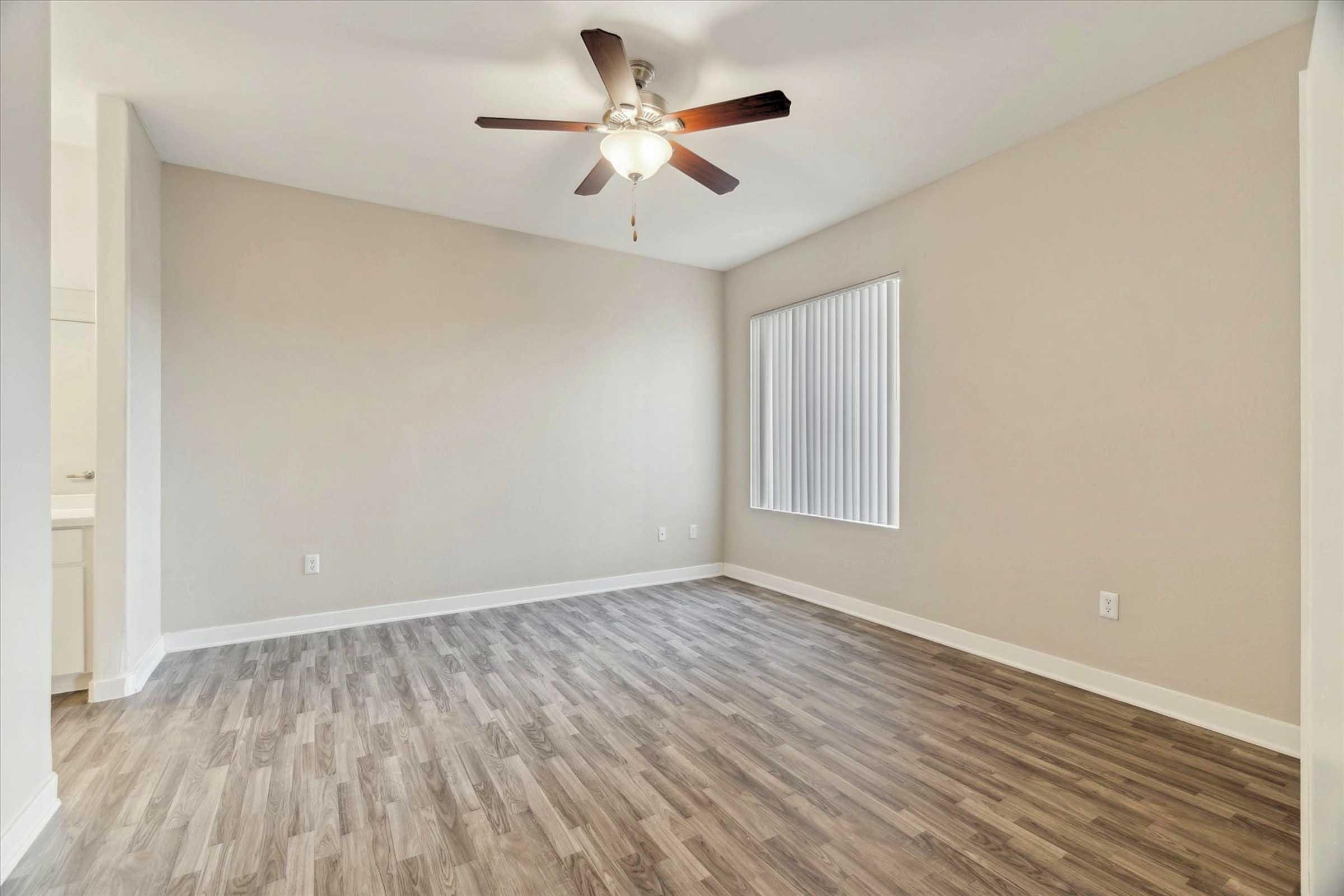 A spacious, empty room with light gray walls and a ceiling fan. The floor is covered in light wood-like laminate. There is a large window with vertical blinds allowing natural light in. The room is uncluttered and ready for furnishings, creating a bright and open atmosphere.