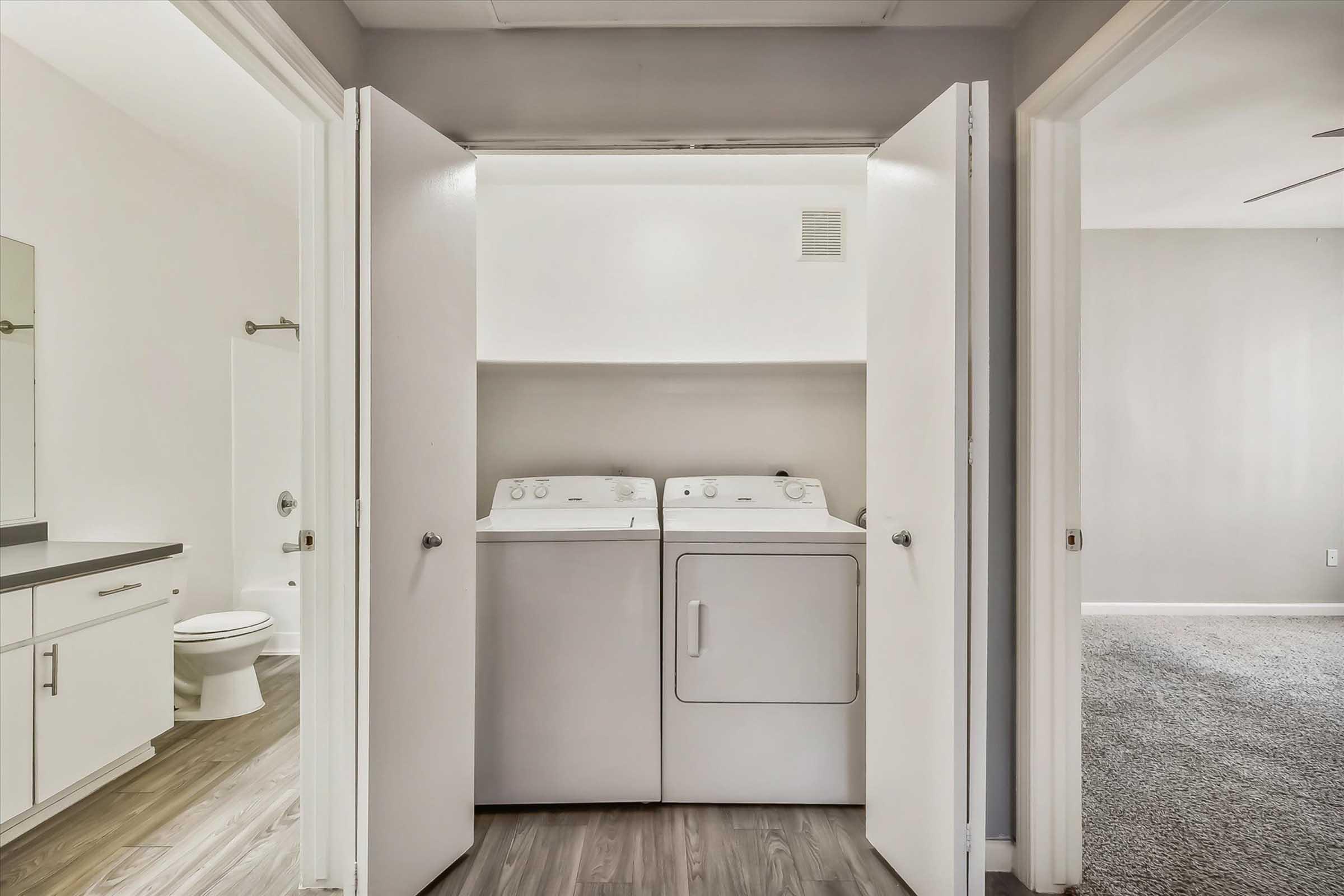 A laundry area featuring a washer and dryer, positioned between two open white doors. In the background, a bathroom with a toilet is visible, along with a carpeted living space to the right. The walls are painted in neutral colors, creating a bright and clean atmosphere.