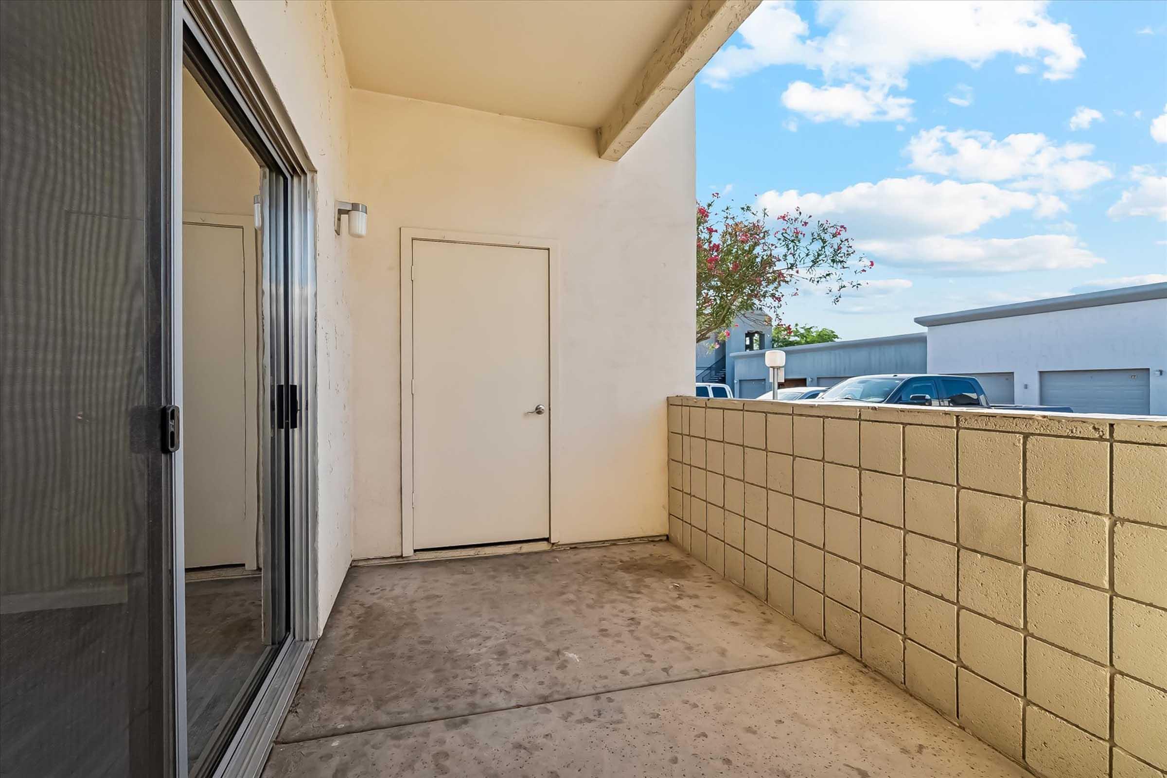 A small outdoor balcony space with a concrete floor and a beige stucco wall. A sliding glass door leads into an interior space, and there is a plain white door on the wall. The area is bright with blue skies and scattered clouds visible in the background.