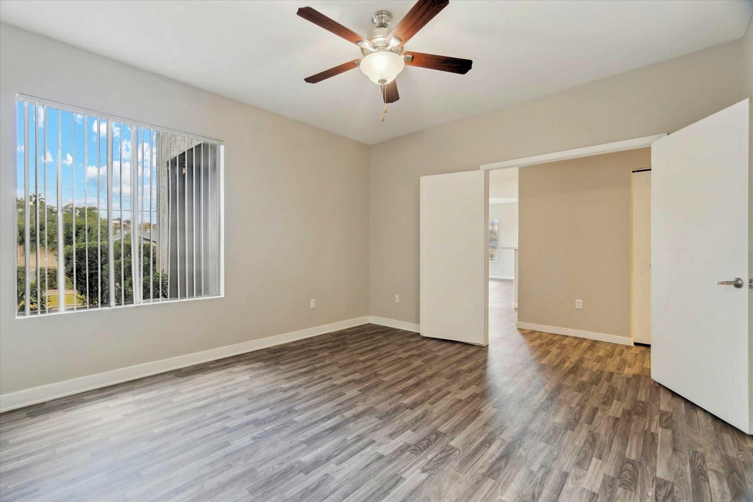 A spacious, well-lit room featuring wooden flooring, a ceiling fan, and large windows with vertical blinds. The room has light-colored walls and a doorway leading to another area, creating an open and airy atmosphere.