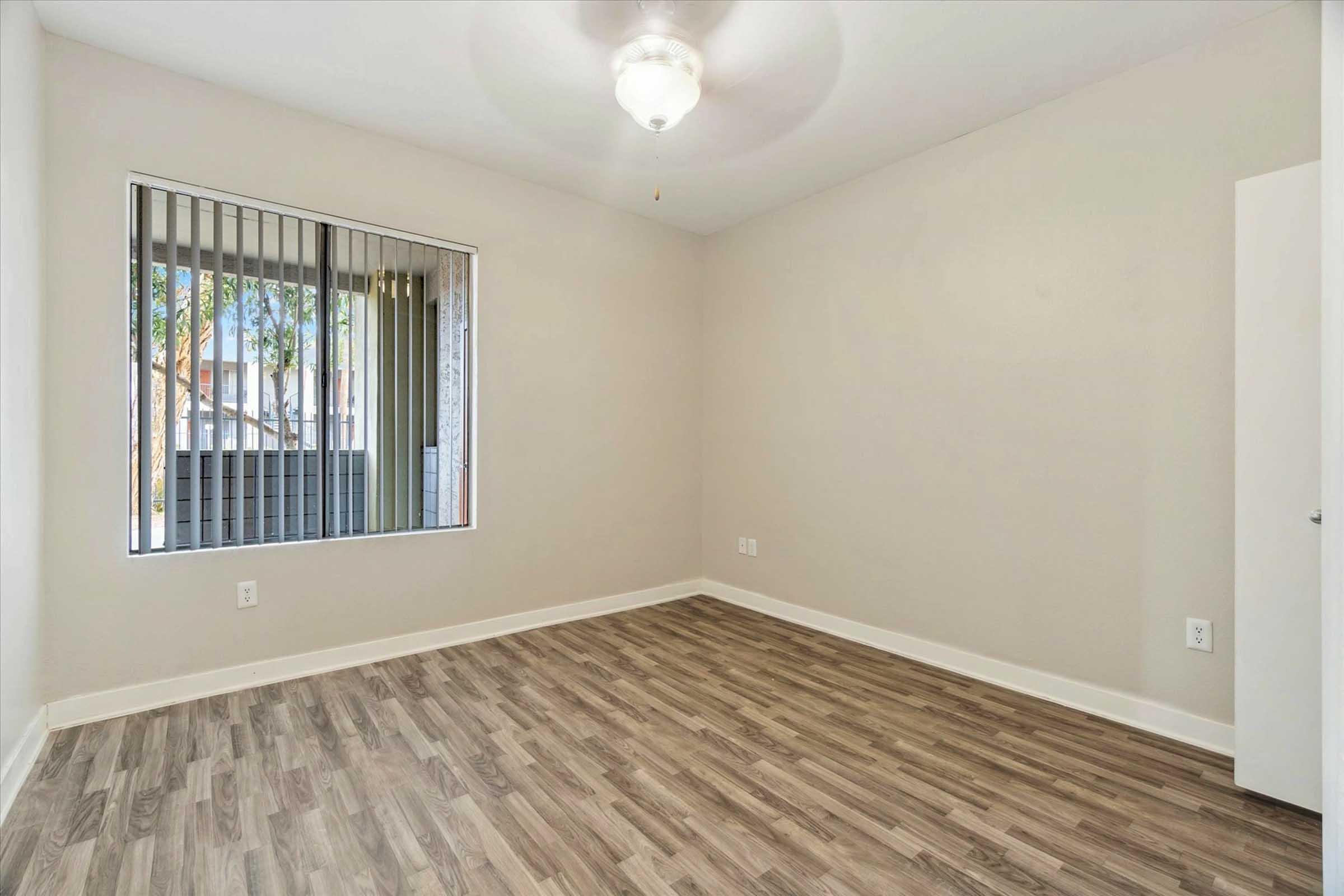A spacious, empty room with light-colored walls and a ceiling fan. It features a large window with vertical blinds, allowing natural light to enter. The floor is covered in light wood laminate, and the room has a simple, modern design. Ideal for personal customization.