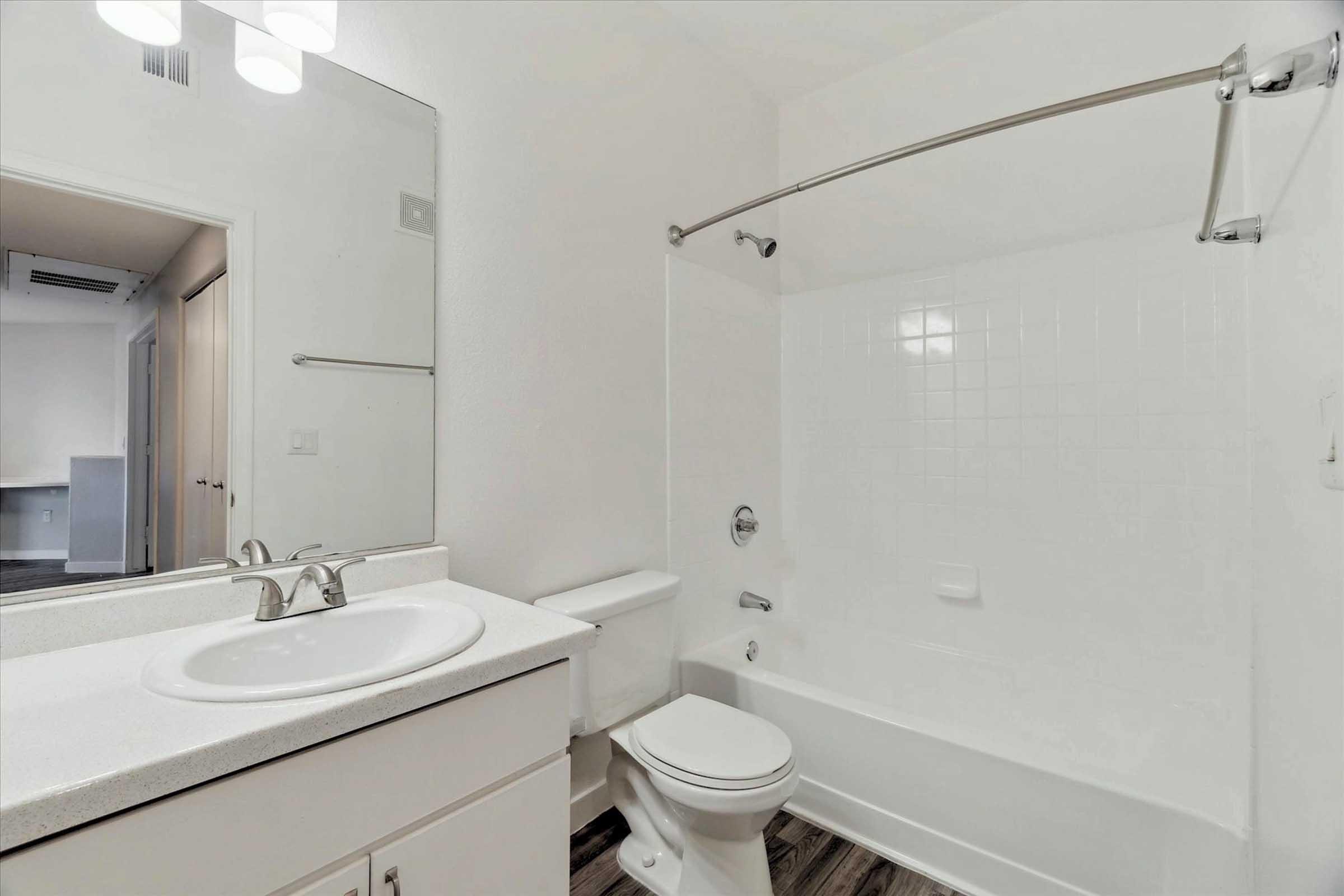 A clean bathroom featuring a white bathtub with a shower, a sink with a countertop, and a toilet. The walls are white, and the floor has a dark wood-like appearance. There's a mirror above the sink and a light fixture overhead. A doorway leads to another room in the background.