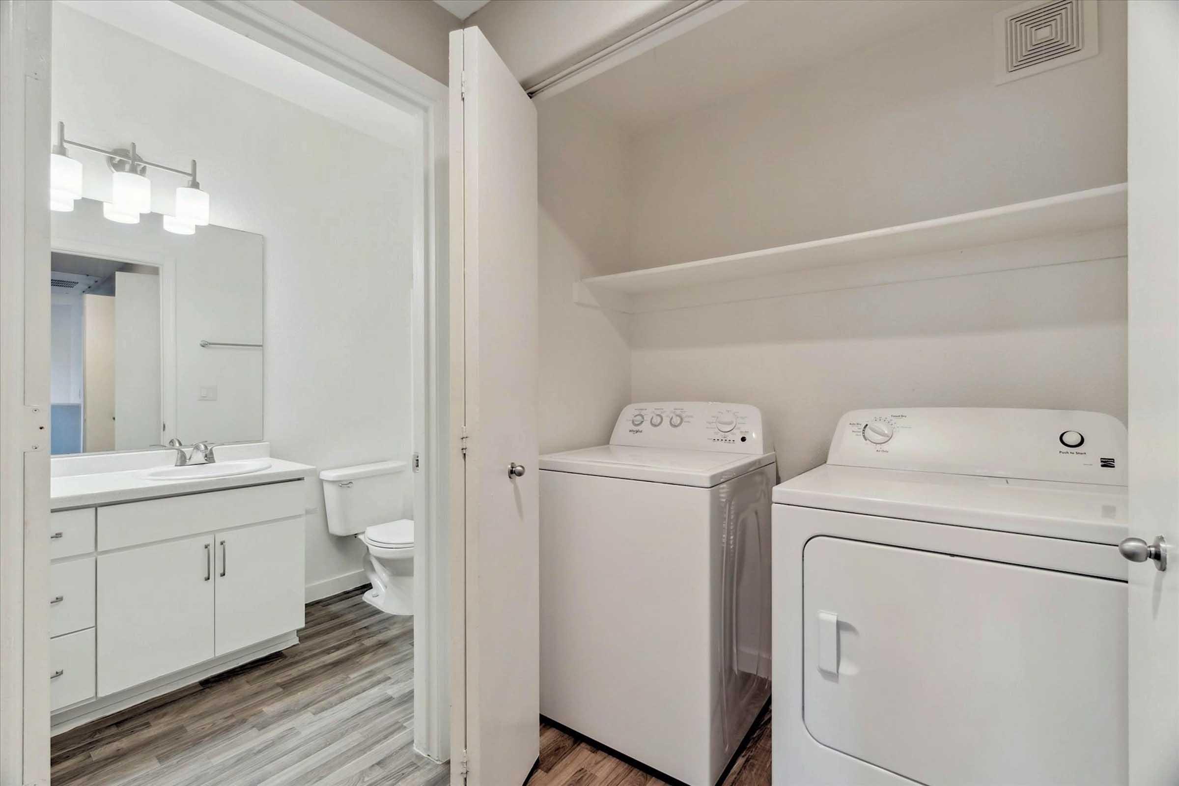 A laundry area featuring a washer and dryer next to a bathroom. The space has white cabinetry and a countertop, with modern light fixtures overhead. The floor has a wood-like finish, and the bathroom includes a toilet and a mirror above the sink.