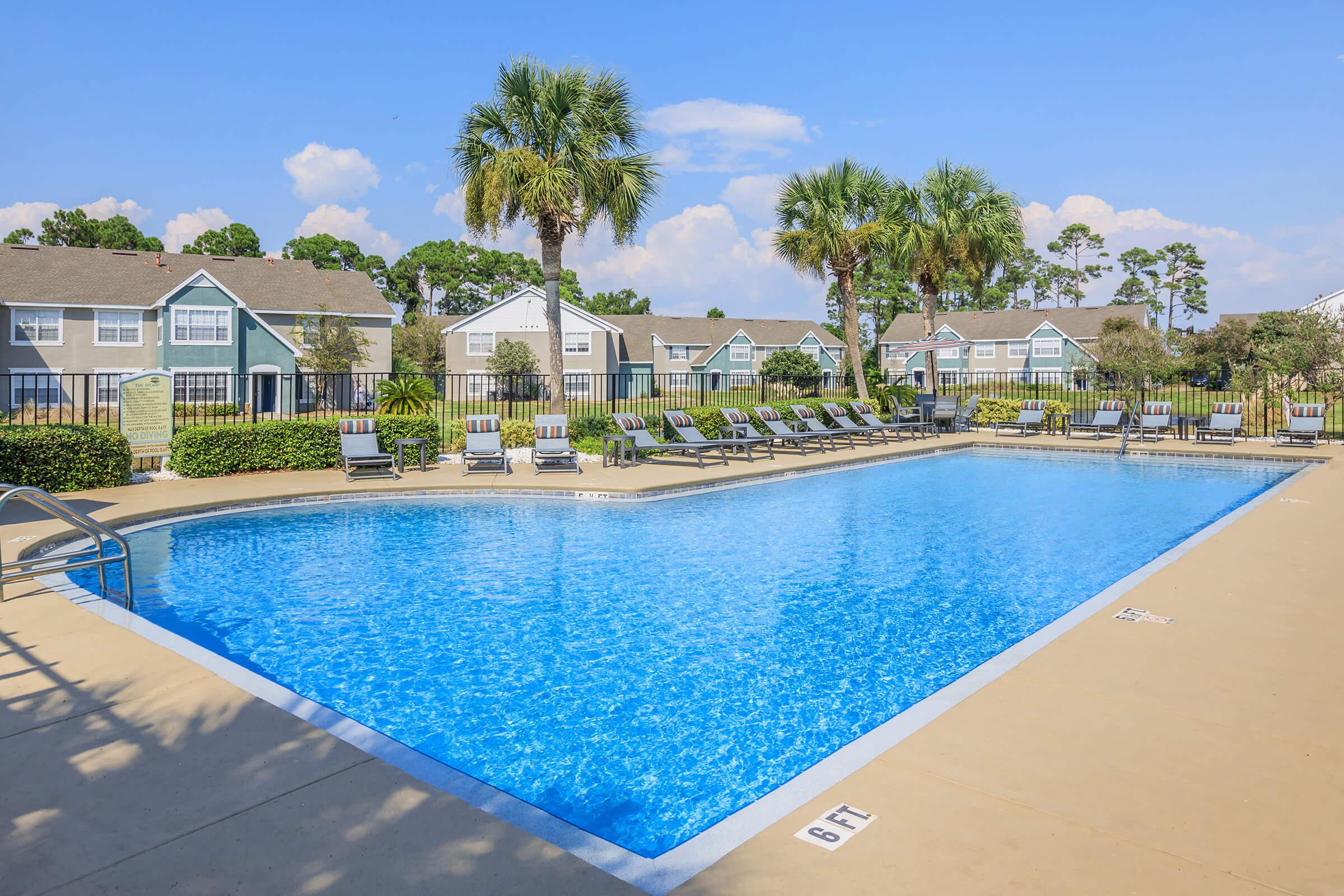 A sparkling swimming pool with a clear blue surface, surrounded by lounge chairs and palm trees. In the background, there are well-maintained residential buildings and a bright blue sky with a few clouds. The pool area looks inviting and relaxing.