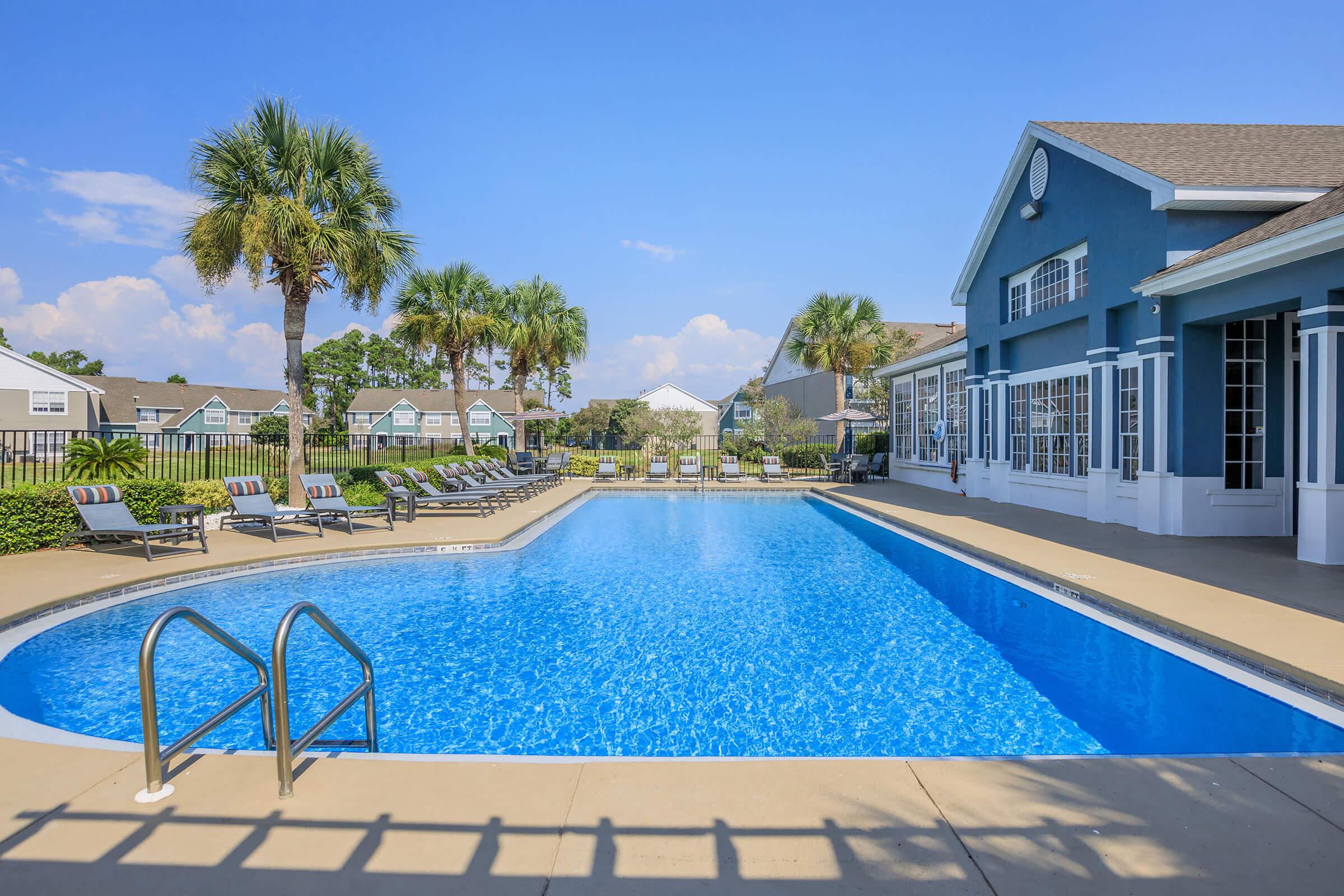 A spacious outdoor pool with clear blue water, surrounded by palm trees and lounge chairs. In the background, there are residential buildings and a clear blue sky. The pool area is well-maintained and inviting, perfect for relaxation and leisure activities.
