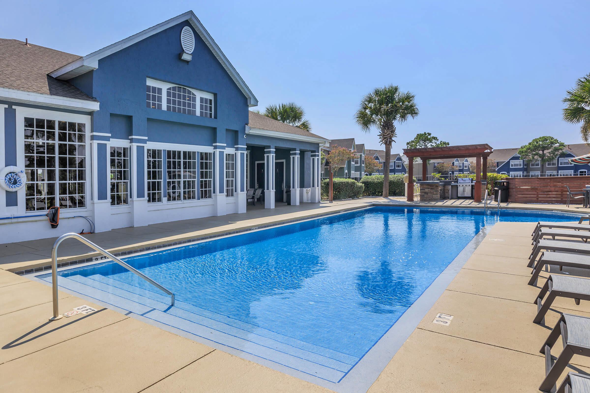 A clear blue swimming pool beside a spacious, well-lit building with large windows and a patio area. Sun loungers line the pool's edge, and palm trees add greenery to the scene. The sky is bright and clear, creating a warm, inviting atmosphere.