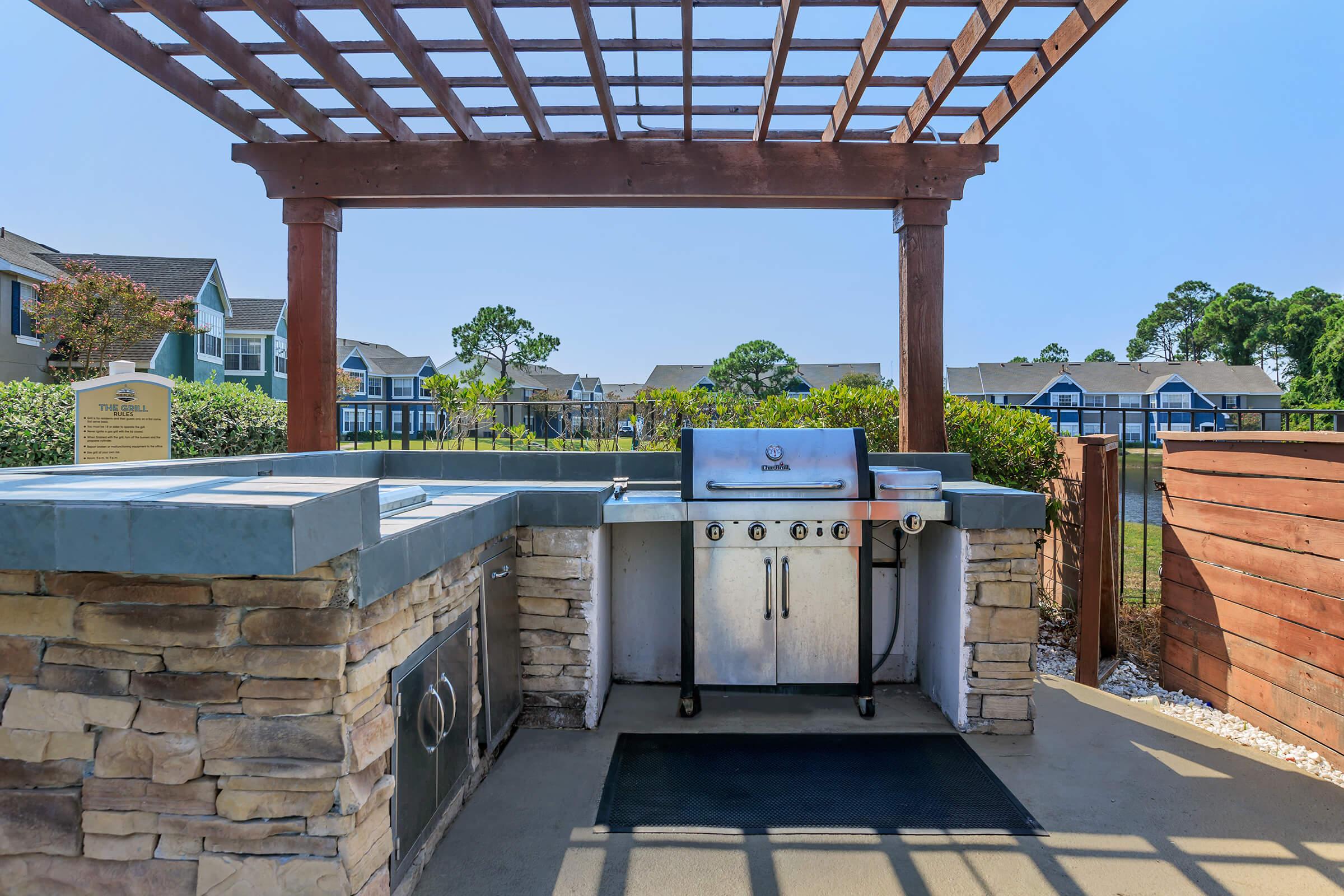 Outdoor barbecue area featuring a stainless steel grill under a wooden pergola. The countertop is made of stone, and there is a side prep station. In the background, there are apartment buildings and lush greenery, with a clear blue sky overhead. Ideal for outdoor cooking and gatherings.