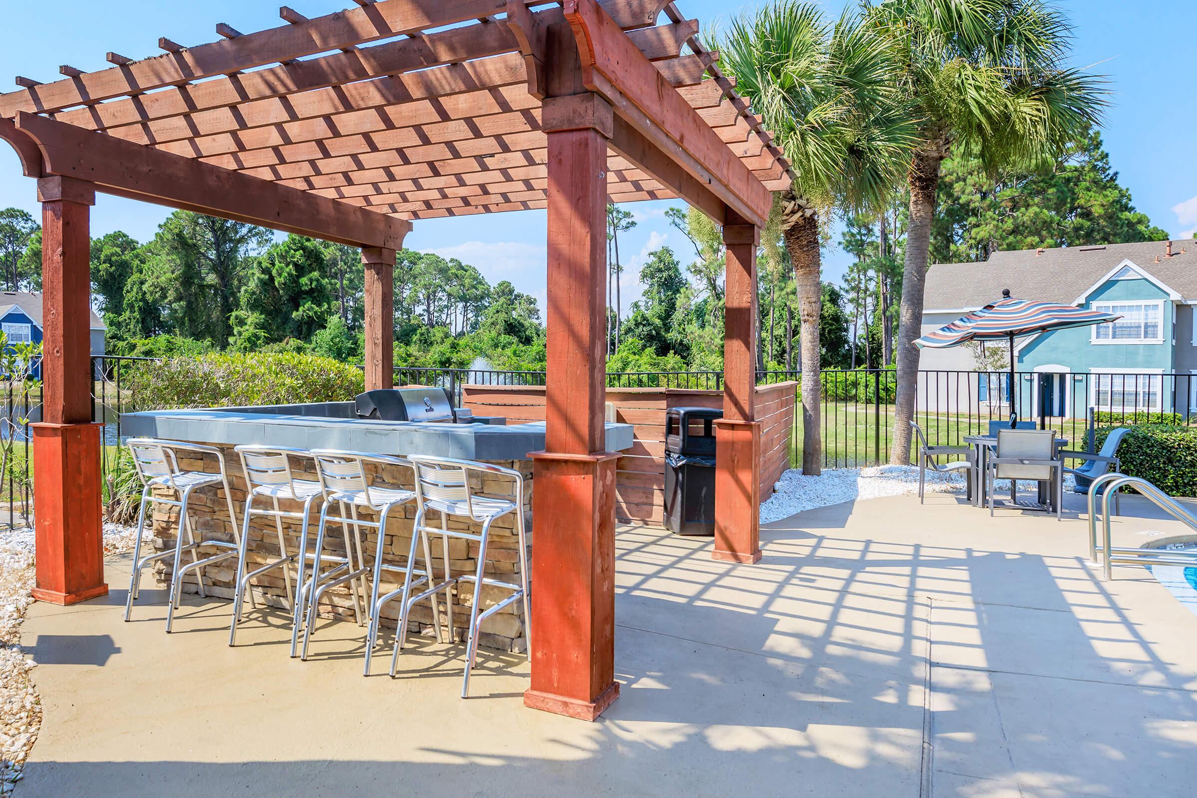 Outdoor patio area featuring a pergola with bar-style seating, surrounded by lush greenery. A swimming pool is visible nearby, with lounge chairs and an umbrella. The setting is bright and inviting, ideal for relaxation and socializing.