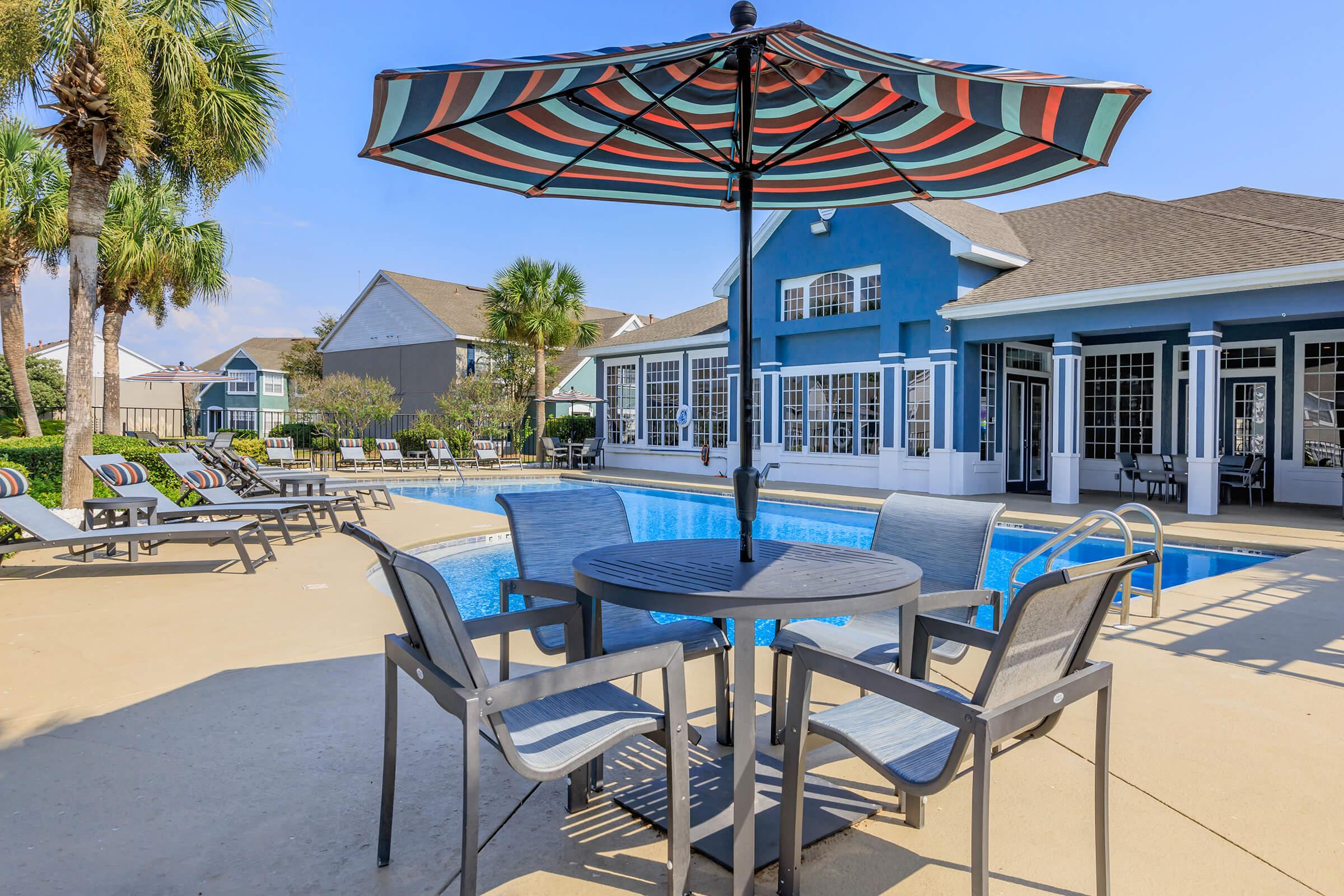 A sunny poolside area featuring a blue pool surrounded by lounge chairs and a shaded table with an umbrella. The backdrop includes a modern building with large windows and palm trees, creating a relaxing atmosphere perfect for leisure and socializing.