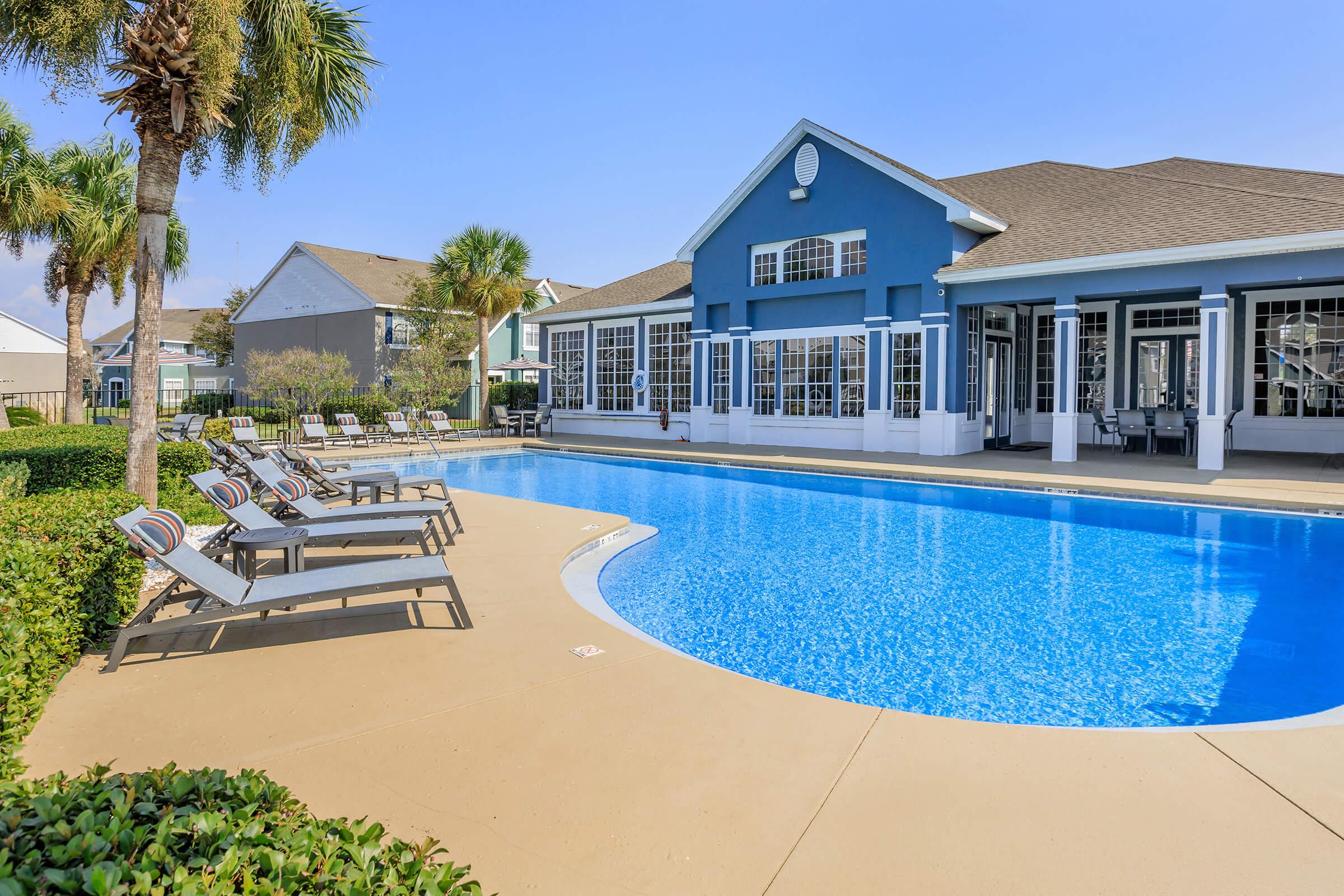 A clear blue swimming pool surrounded by lounge chairs, with a modern building in the background. Lush greenery and palm trees add to the inviting atmosphere on a sunny day. The scene conveys a relaxing outdoor space ideal for leisure and enjoyment.