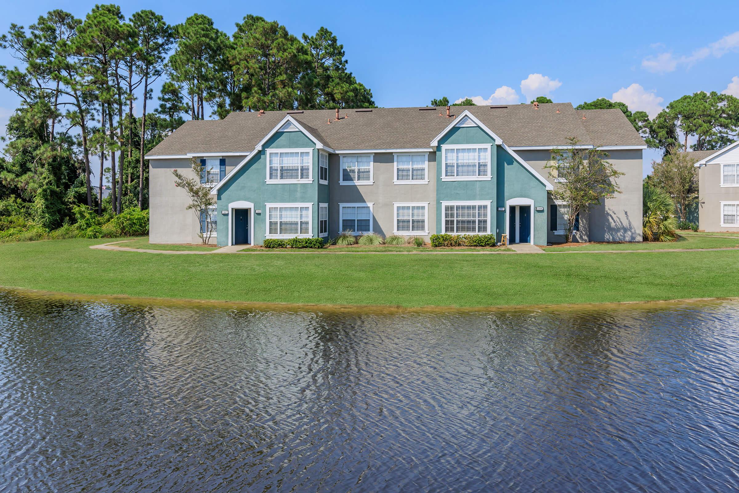 Two-story residential building with a light gray and green exterior, situated by a pond. Lush green lawn surrounds the building, and tall trees in the background provide a natural setting under a clear blue sky. Reflections of the building and trees can be seen in the water.