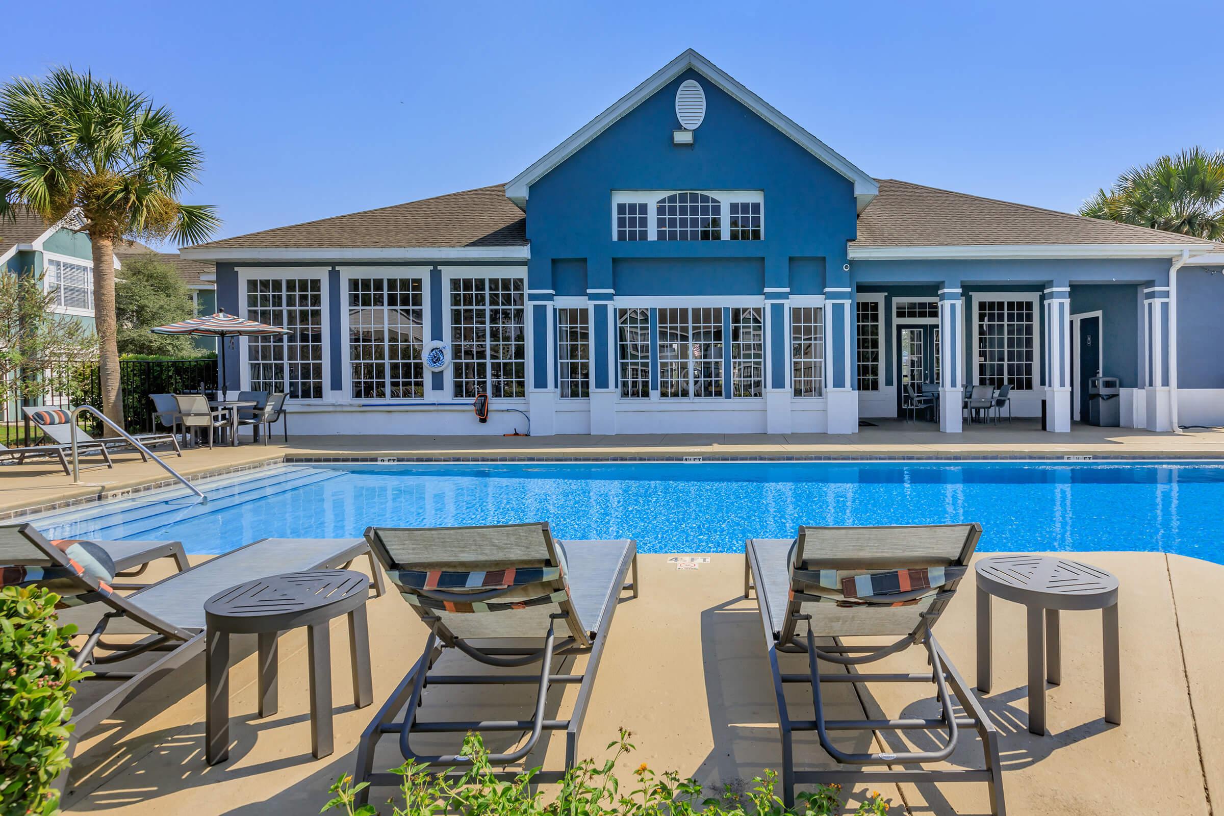 A swimming pool surrounded by lounge chairs, with a blue building featuring large windows in the background. Palm trees are visible, and the sky is clear and sunny. The pool's surface reflects the serene environment, creating a relaxing atmosphere.
