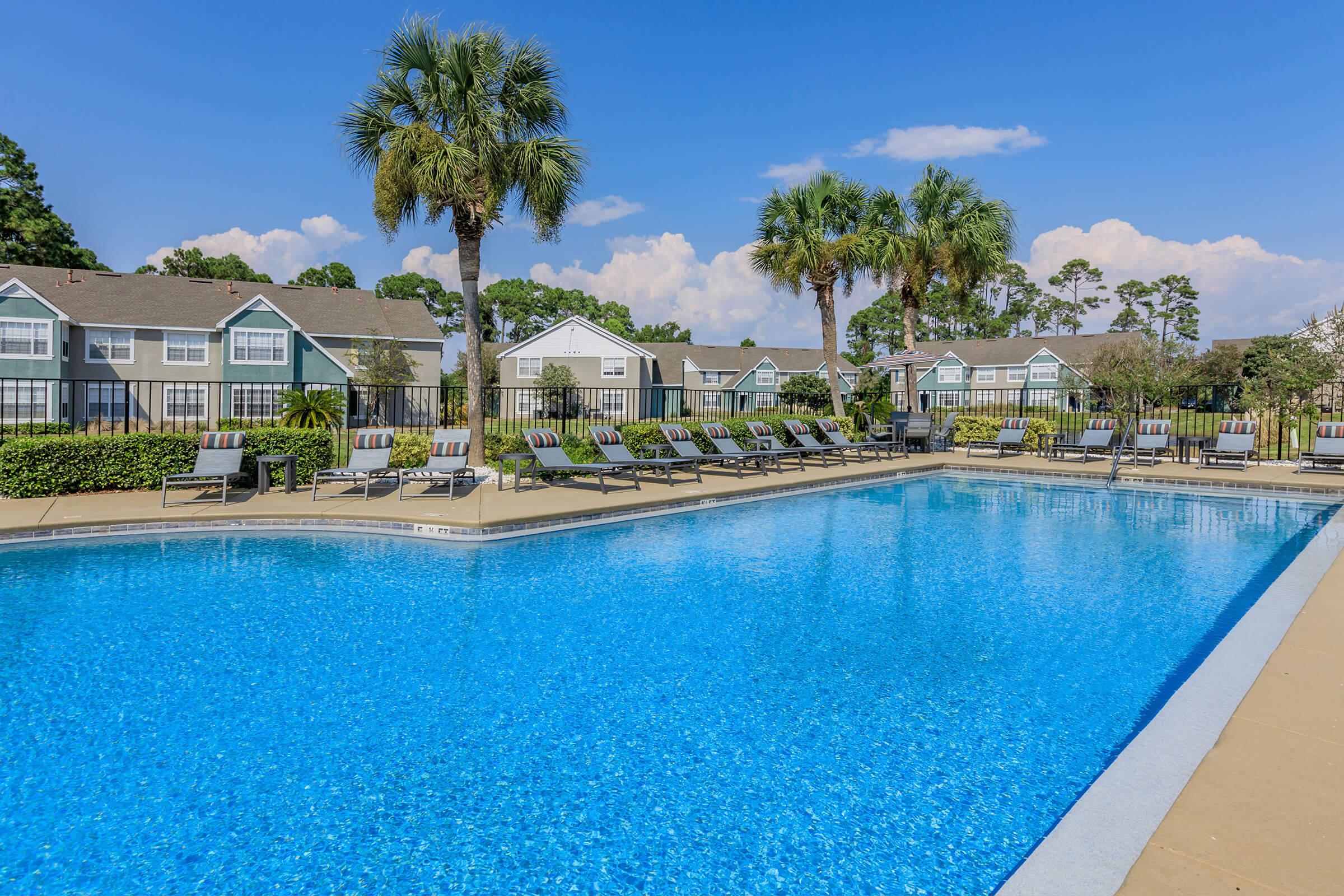 A clear blue swimming pool surrounded by lounge chairs, with lush greenery and palm trees in the background. Residential buildings are visible on the far side, under a bright blue sky with a few fluffy clouds. The scene conveys a relaxed, inviting atmosphere ideal for leisure and relaxation.