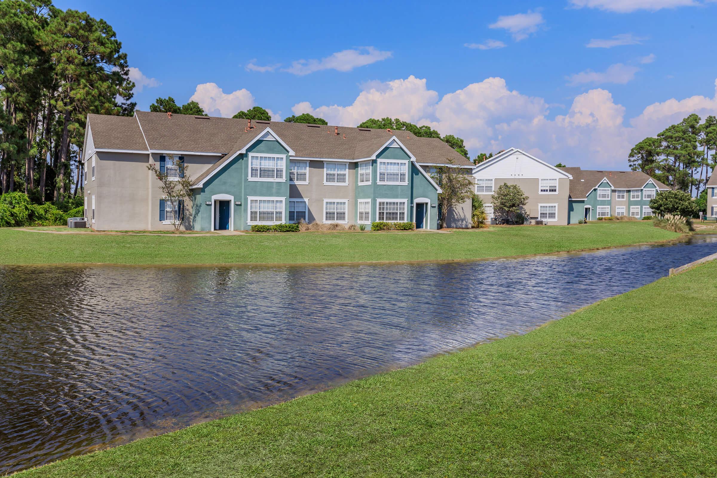 A scenic view of two multi-unit residential buildings with green and blue exteriors, situated beside a calm pond. Lush green grass surrounds the water, and the sky features scattered clouds, creating a peaceful suburban atmosphere.