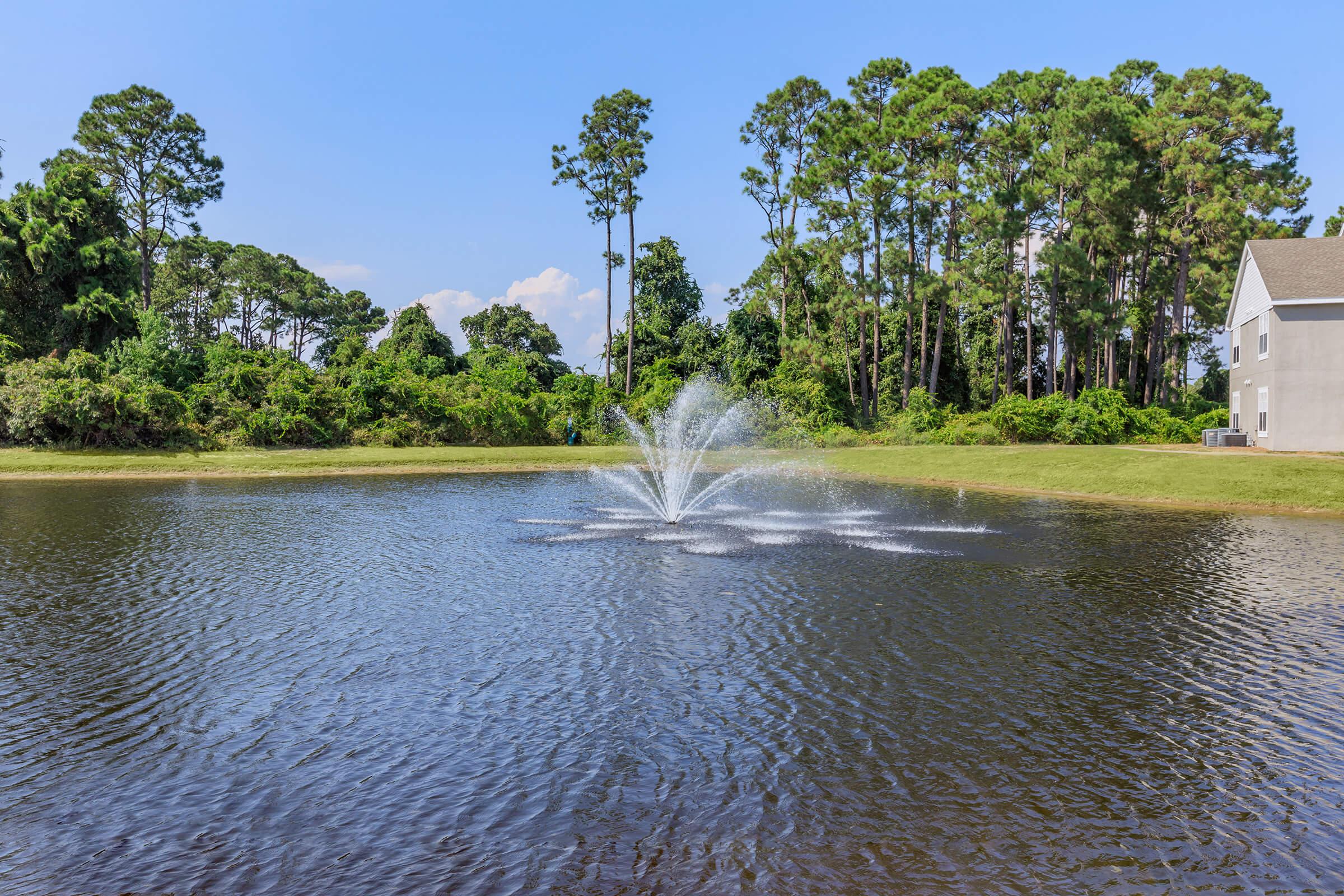 A tranquil pond surrounded by lush trees and greenery, featuring a fountain that sprays water into the air, creating a serene atmosphere under a clear blue sky.