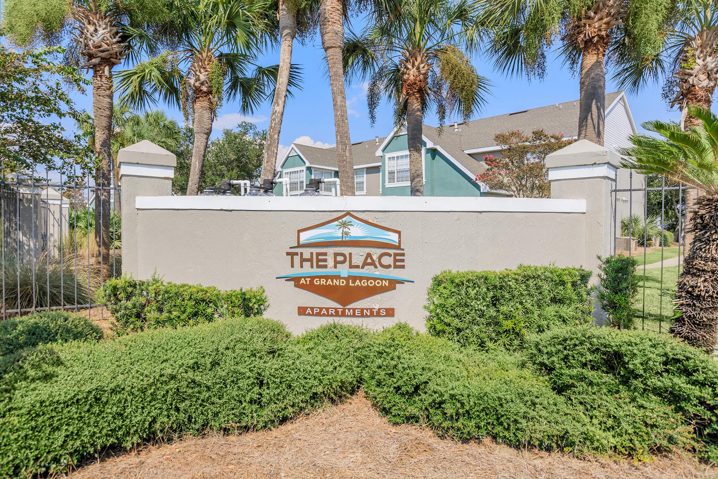 Sign for "The Place at Grand Lagoon Apartments" displayed prominently, surrounded by landscaped greenery and palm trees. The setting features a well-kept garden area, with residential buildings visible in the background.
