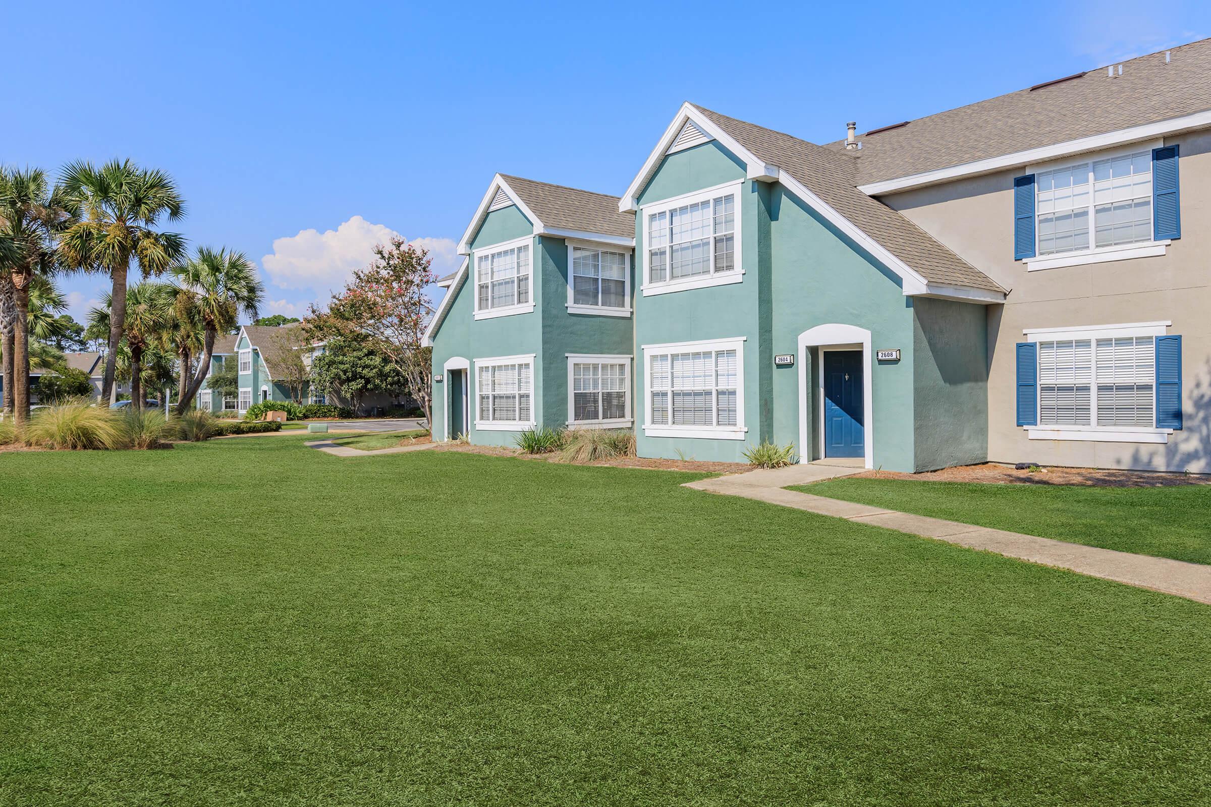 Two-story apartment buildings with green and gray siding, surrounded by a well-maintained lawn. The sky is clear and blue, with some trees and shrubs in the background, creating a serene residential atmosphere. Pathways lead to the entrances of the apartments.