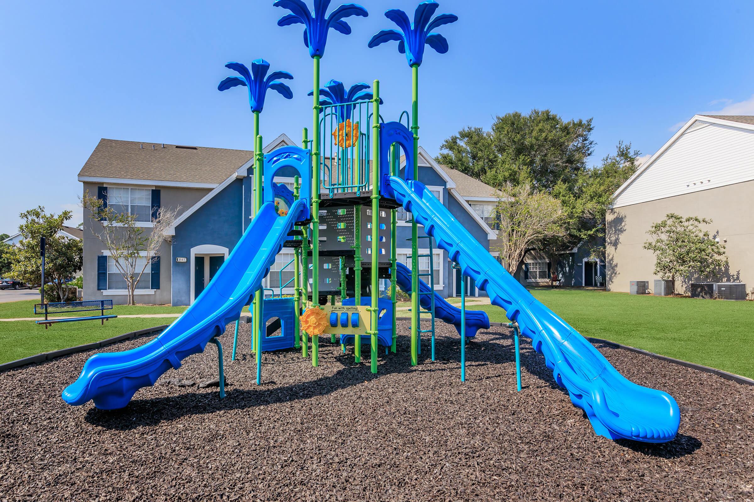 Brightly colored playground featuring a two-story blue slide structure surrounded by palm tree designs. The structure includes two twisting slides and is set on a bed of dark mulch, with green grass and buildings in the background under a clear blue sky.