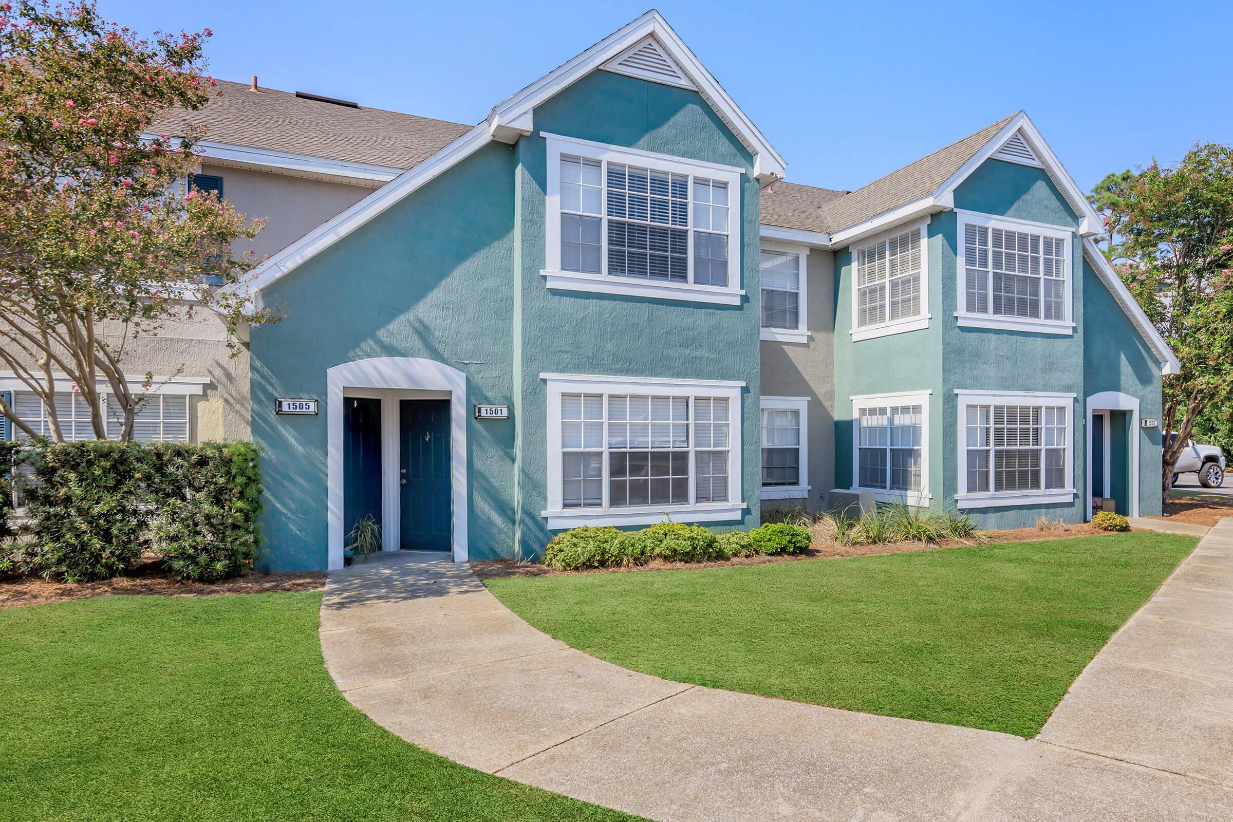A multi-unit residential building featuring two stories with a light blue and gray exterior. The property has several windows, a well-maintained lawn, and a paved walkway leading to the entrance. Bright sunlight illuminates the scene, enhancing the vibrant colors of the building.