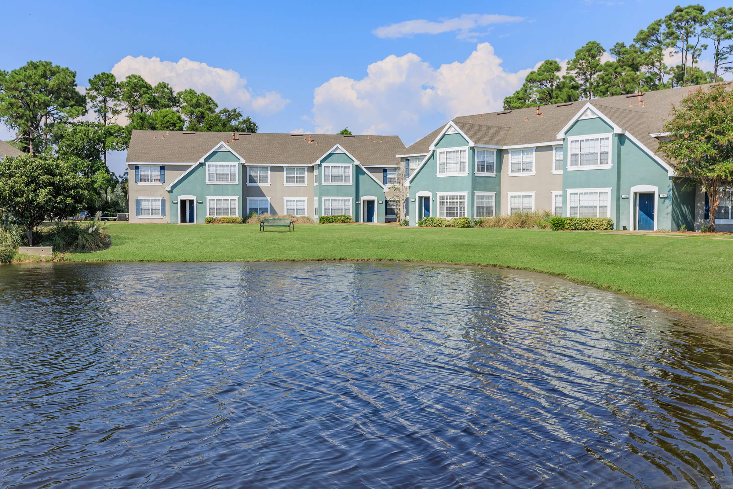 A scenic view of a residential area featuring blue and white apartment buildings beside a calm pond. Lush green grass and trees surround the area, with a clear blue sky and a few clouds overhead. The tranquil setting suggests a peaceful community atmosphere.