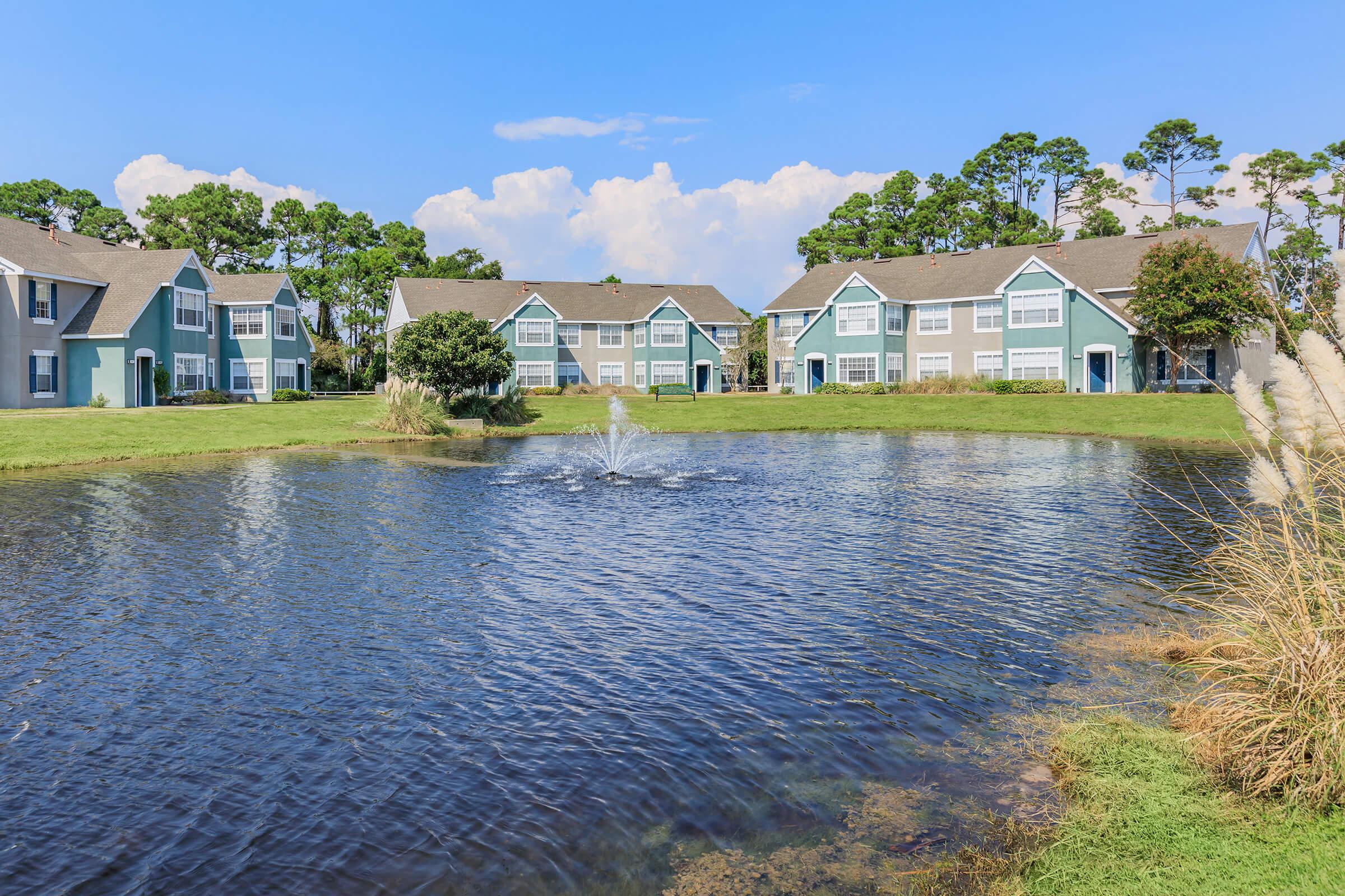 A serene landscape featuring a pond with a fountain, surrounded by green grass and clustered apartment buildings with blue and gray exteriors. In the background, there are trees under a clear blue sky, creating a tranquil residential setting.