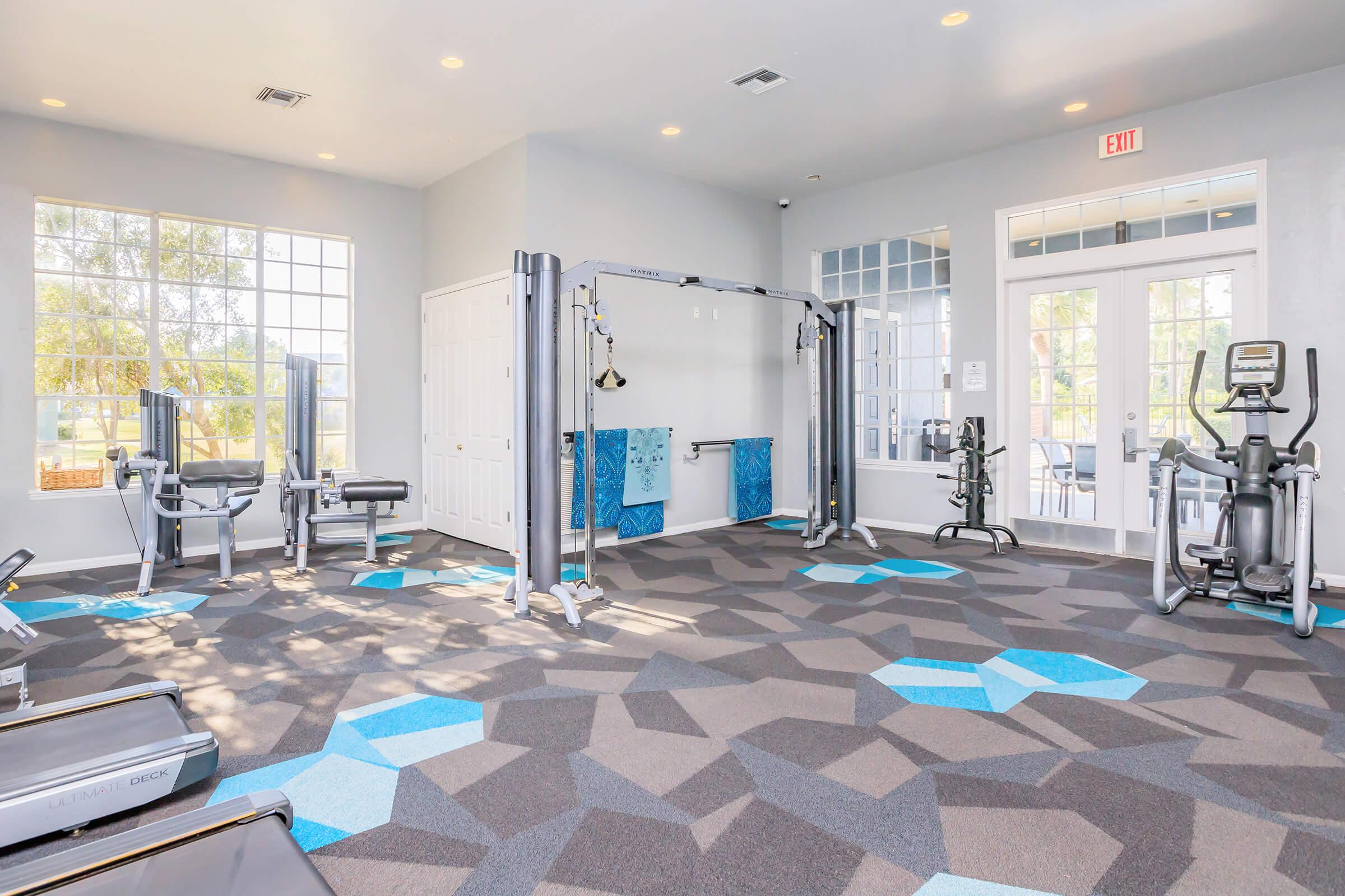 A bright, modern fitness center featuring multiple workout machines, including cable and elliptical equipment. The room has large windows allowing natural light, with blue and gray hexagonal carpet tiles. There are towels on a rack and a glass door leading outside, creating an inviting workout space.