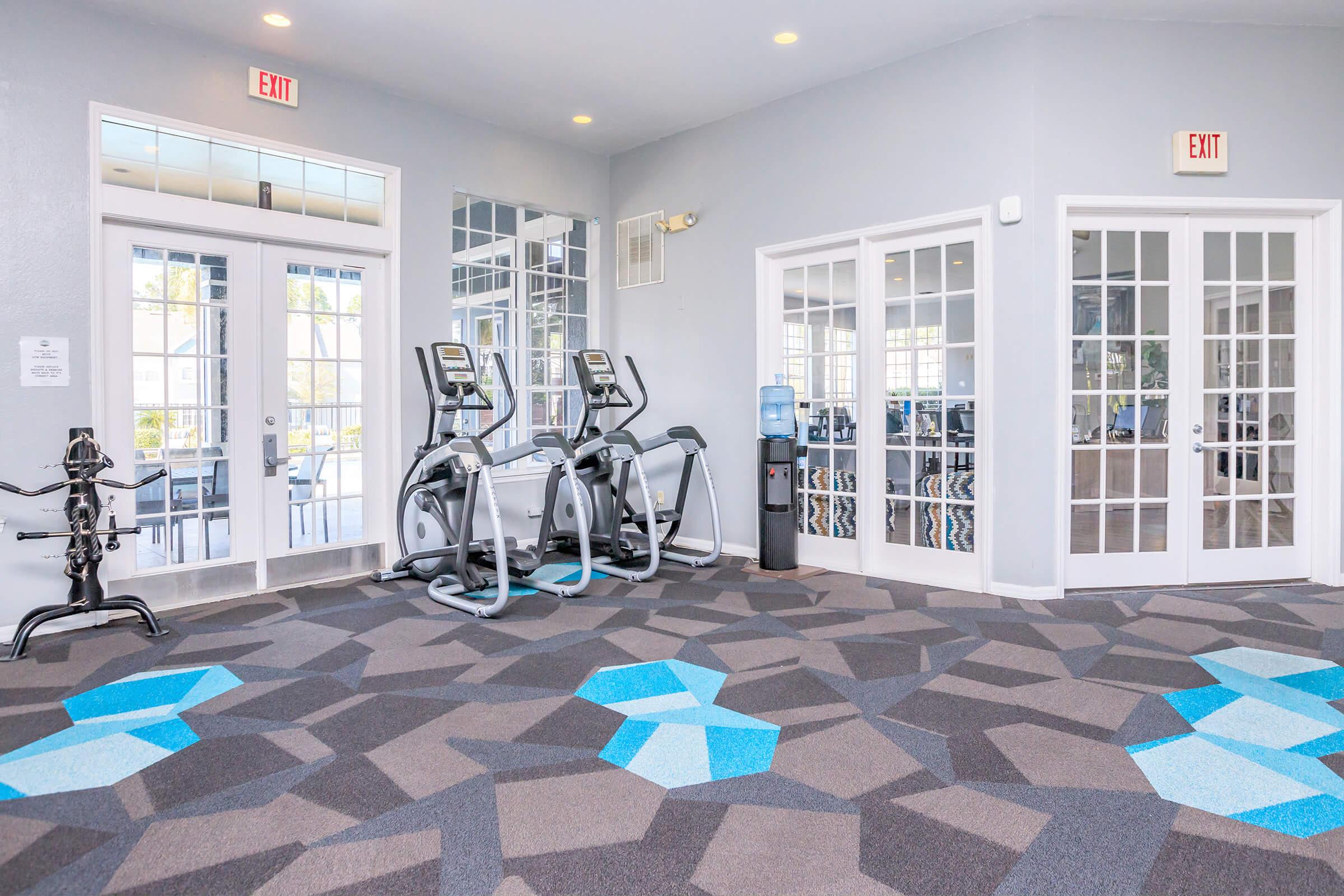 A spacious fitness center featuring gray walls and an eye-catching carpet design in blue and gray. The room contains two elliptical machines, a water cooler, and large windows letting in natural light. Mirrors reflect the workout space, enhancing the open feel. Multiple exits lead to outdoor areas.