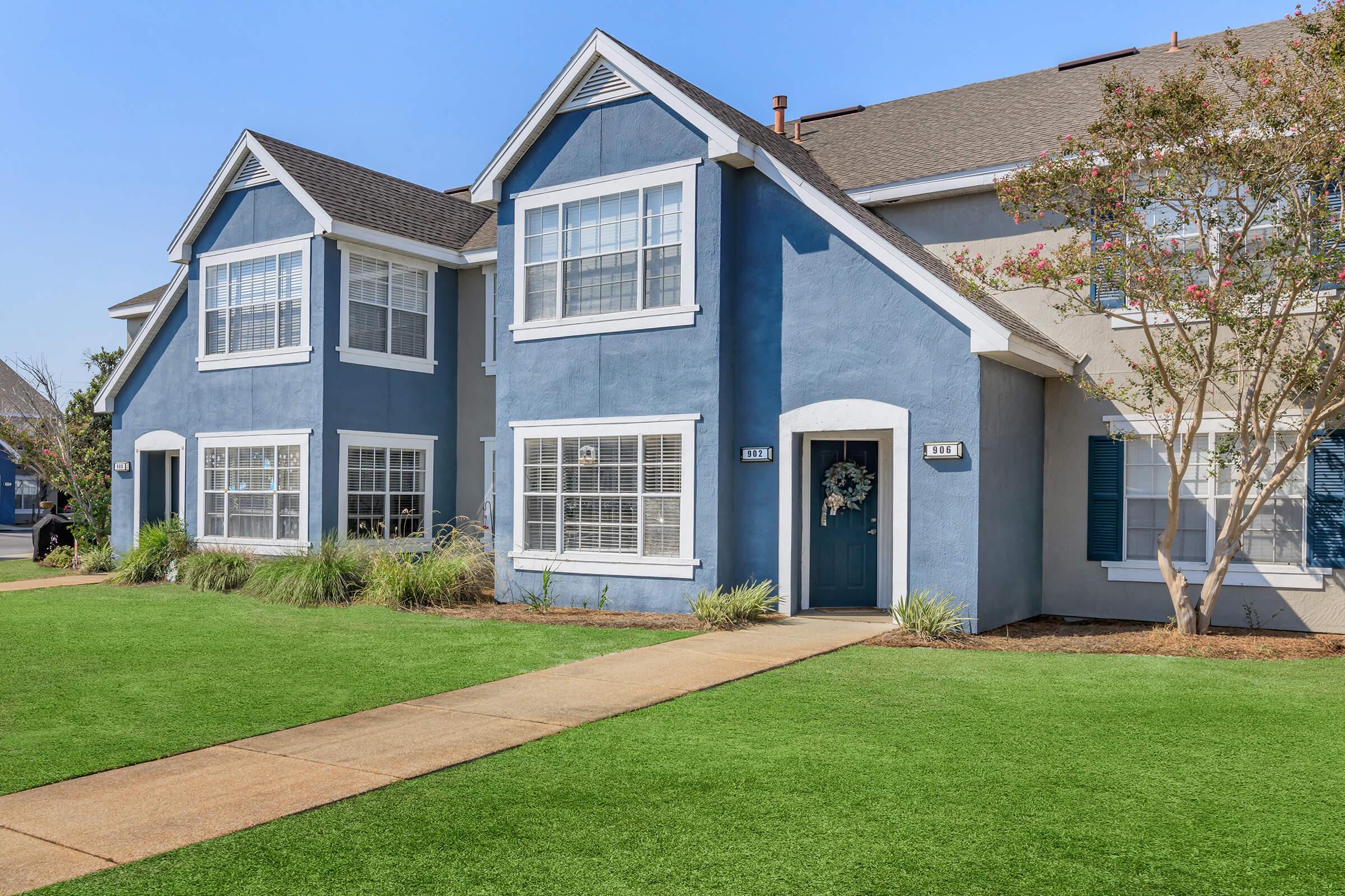 A well-maintained suburban apartment complex featuring two connected units. The exteriors are painted in a blue and beige color scheme, with white trim and large windows. Lush green grass surrounds the building, and a concrete pathway leads to the entrance. Trees and shrubs add to the landscaped appearance.