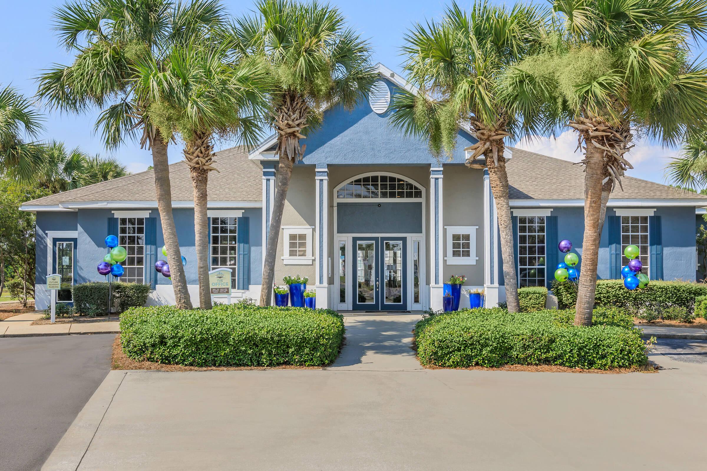 A modern building with blue and gray facades, featuring large front windows and a welcoming entrance. Palm trees and decorative planters surround the entrance, and colorful balloons are tied to the railing, suggesting a festive atmosphere. The driveway is paved, leading up to the entrance.