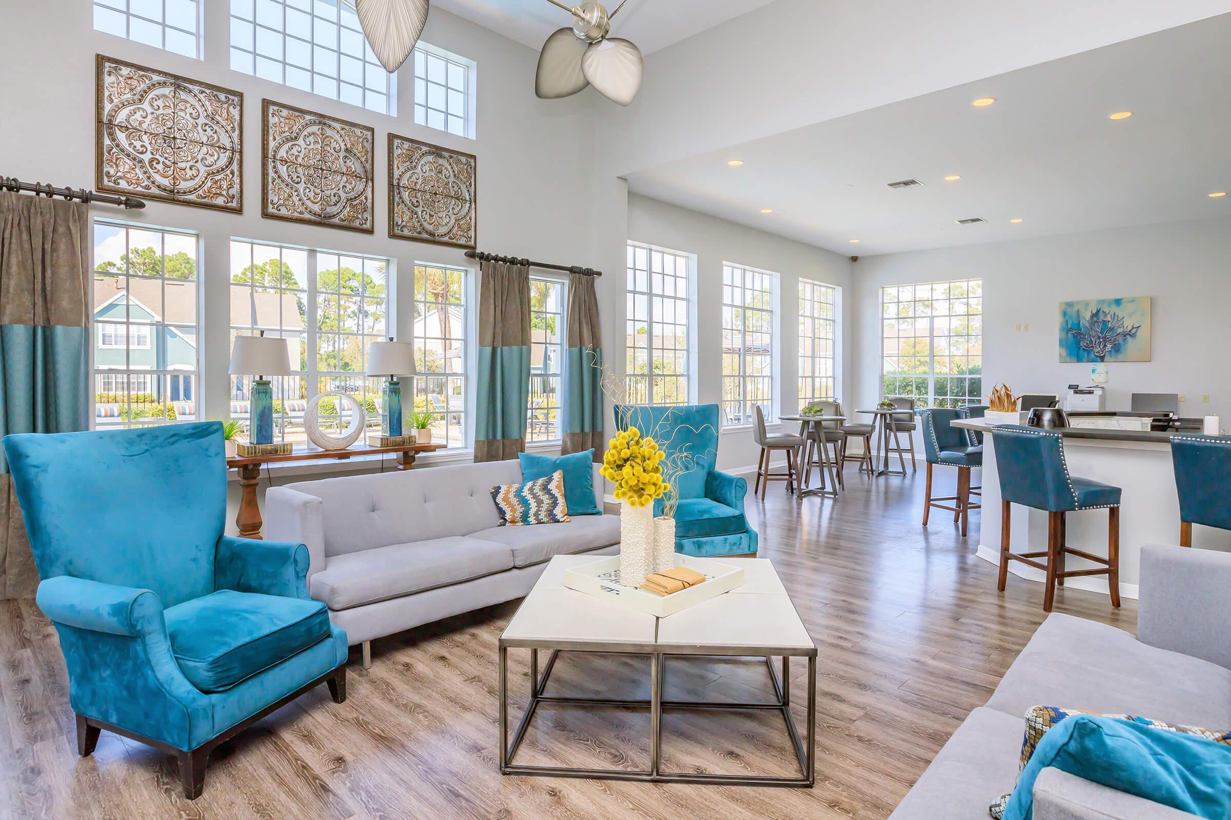 A modern, brightly lit lounge area featuring blue upholstered chairs and gray sofas, decorated with yellow flowers on a coffee table. Large windows allow natural light, and artistic wall decor adds character. The space includes a dining area with stools, creating a welcoming atmosphere for relaxation or socializing.
