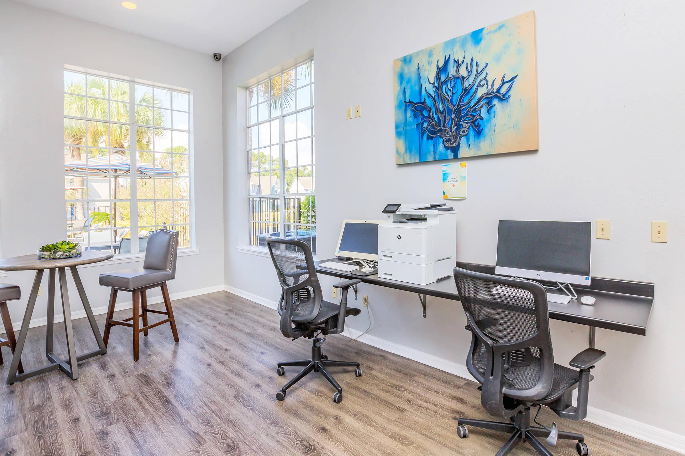 A modern office space featuring two desks with computer monitors, an all-in-one printer, and ergonomic chairs. Large windows allow natural light to brighten the room, and a decorative blue coral artwork hangs on the wall. There are plants on the table, enhancing the workspace's aesthetic.