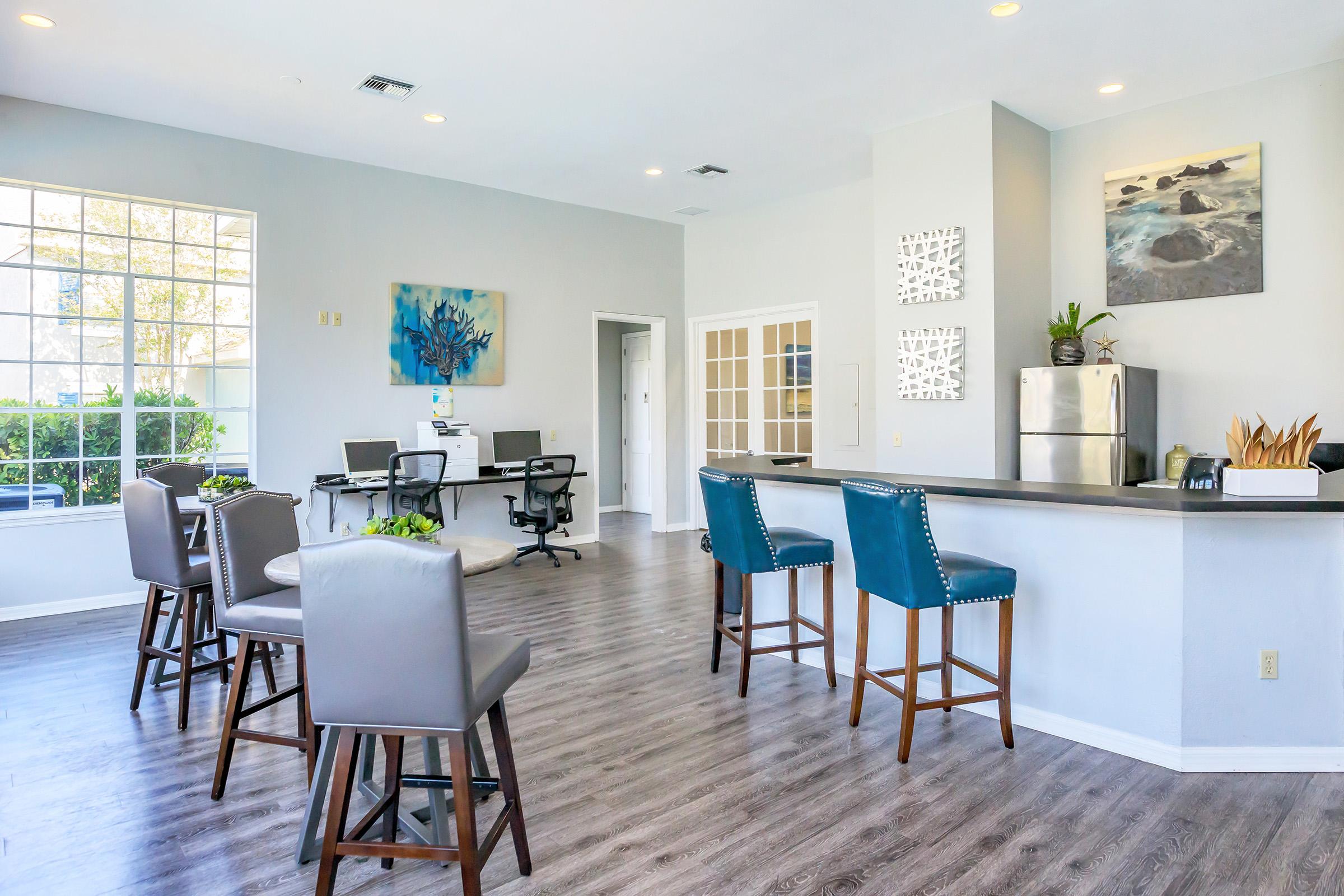 A modern interior space featuring a kitchenette with bar stools, a small dining area, and workstations with computers. Large windows allow natural light to illuminate the room, decorated with plants and artwork on the walls. The floor is wood-like laminate, creating a warm and inviting atmosphere.