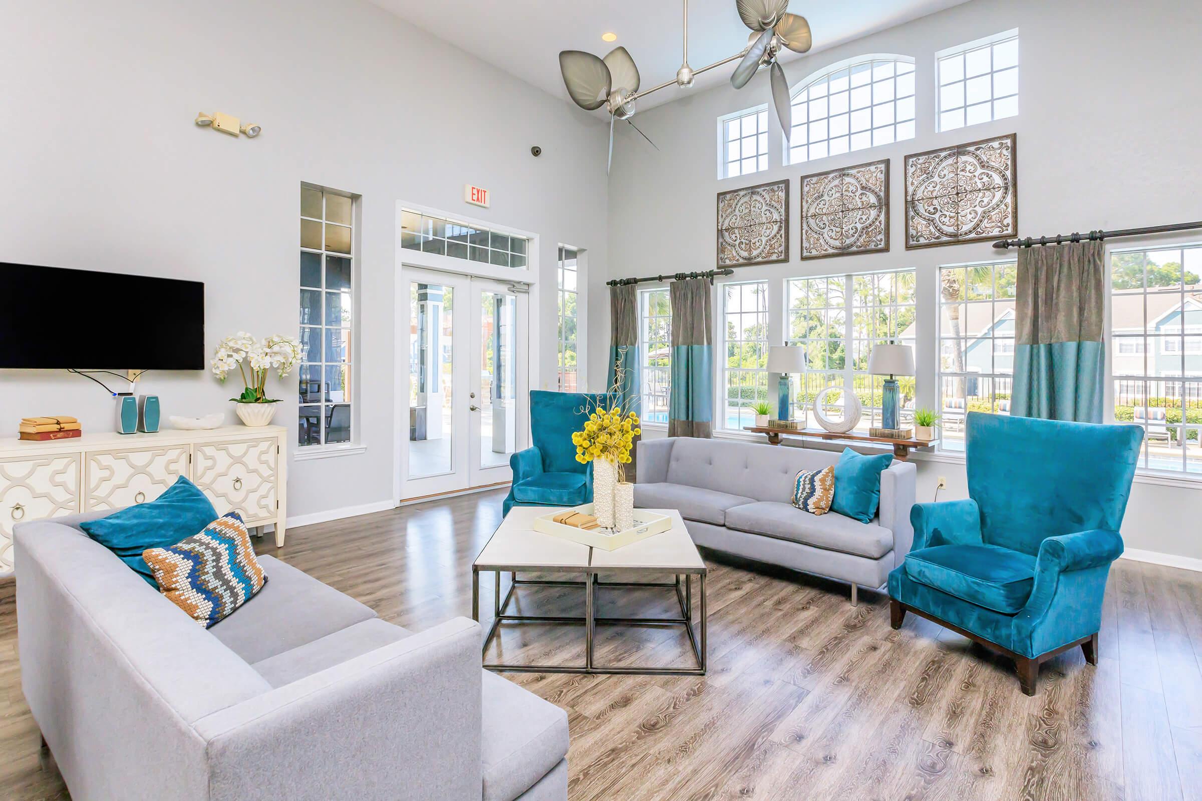 Bright and modern lounge area with large windows, featuring gray sofas and vibrant blue armchairs. A coffee table sits in the center, adorned with a yellow flower arrangement. Decorative wall accents and a TV enhance the inviting atmosphere of the space.