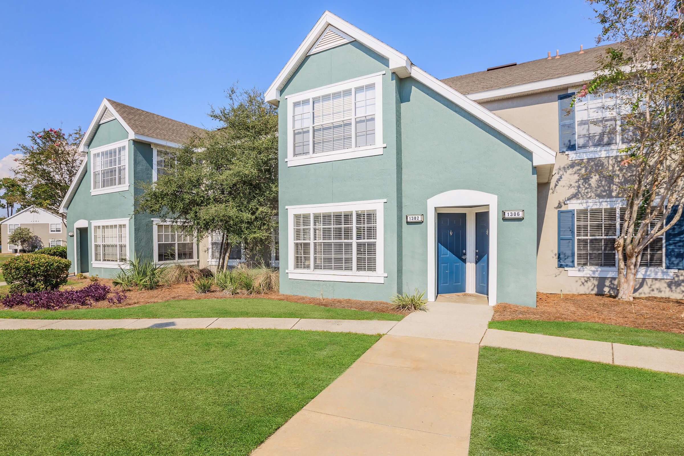 Exterior view of a two-story apartment building featuring green and beige siding. The entrance has a blue door, surrounded by landscaped grass and shrubs. Clear skies with minimal clouds are visible above. The building has multiple windows, and the pathway leads to the entrance.