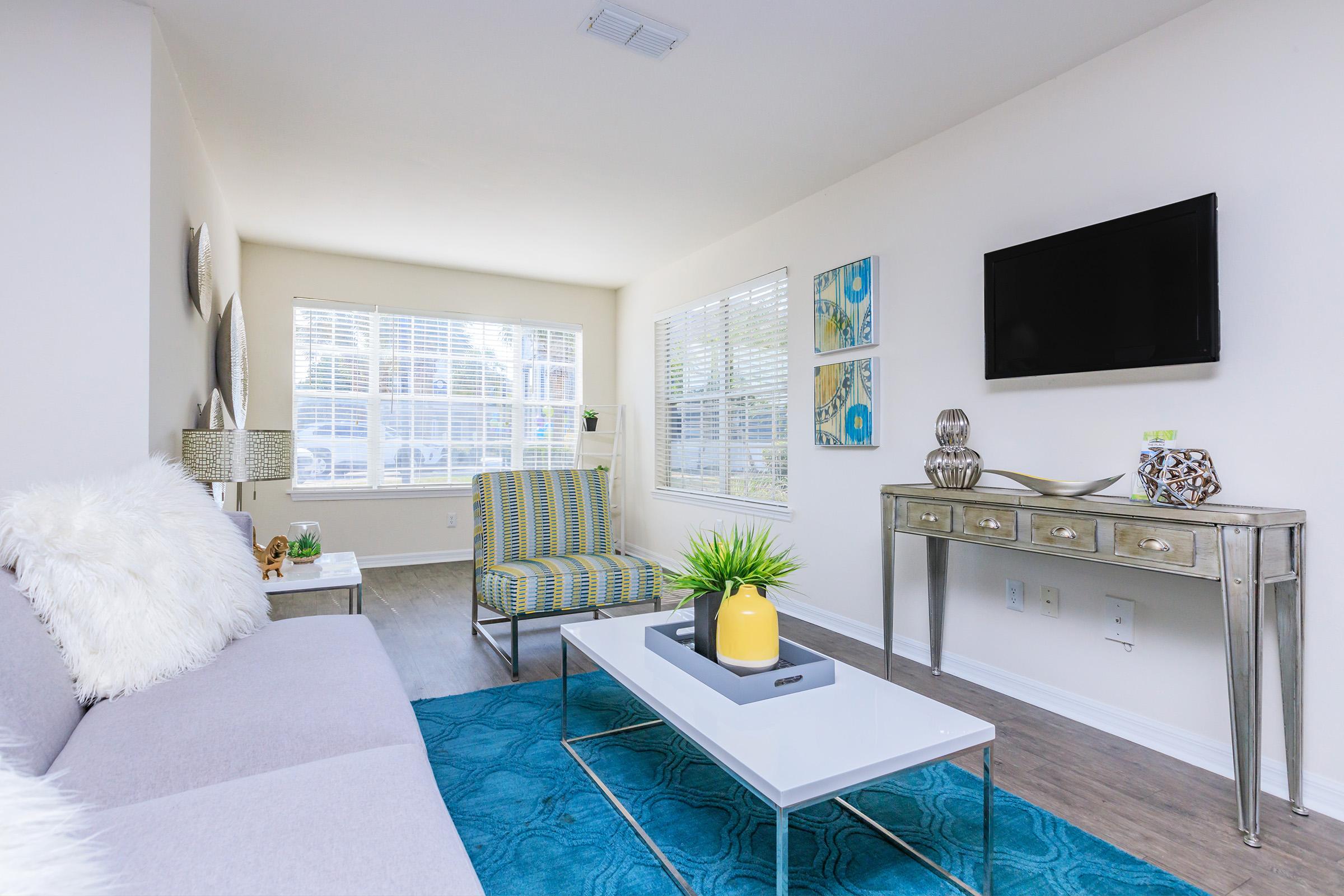 A stylish living room featuring a gray sofa with plush pillows, a striped accent chair, and a coffee table with a decorative plant. The space has large windows allowing natural light, light-colored walls, and modern artwork on display. Wood flooring adds warmth to the contemporary decor.