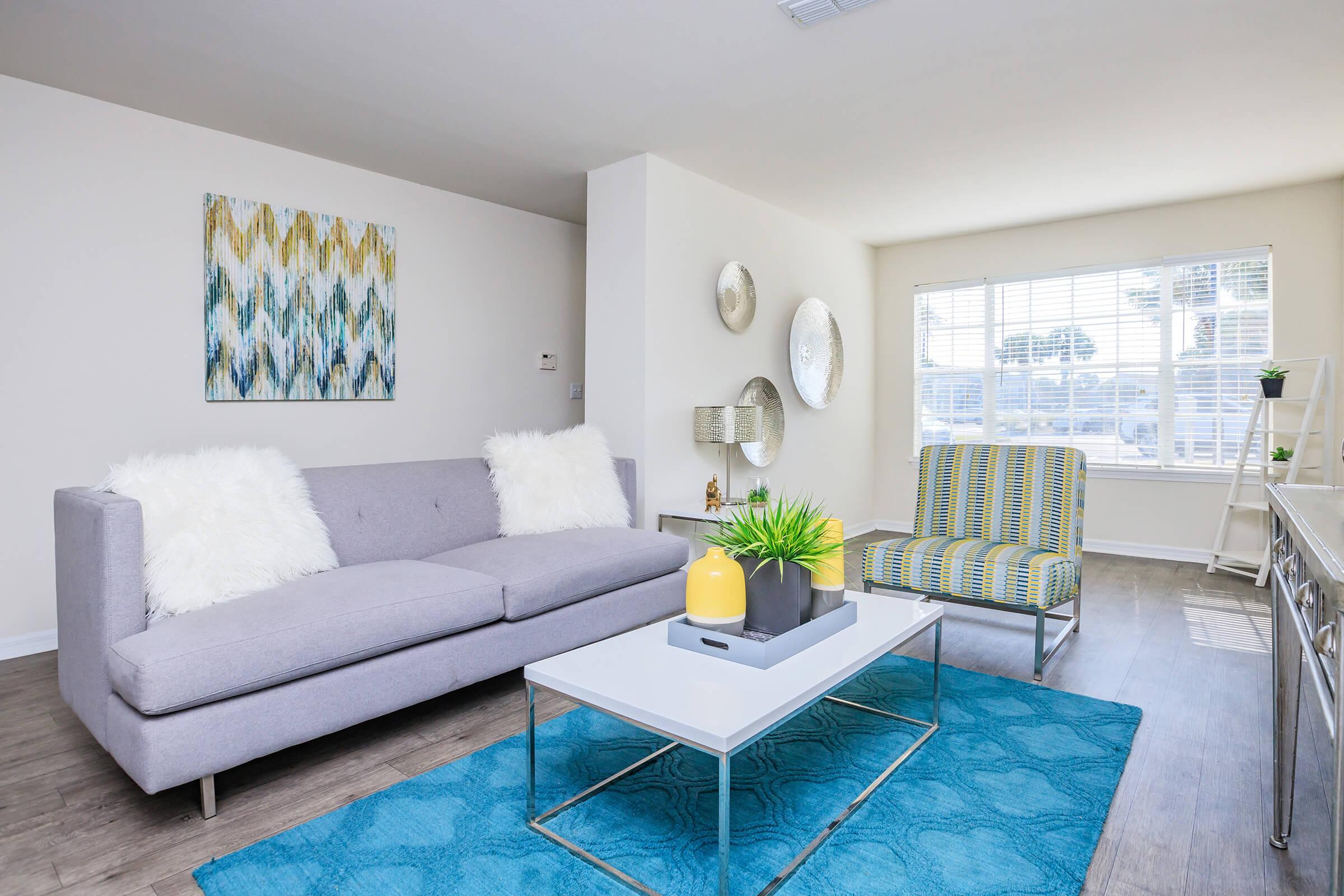 A cozy living room featuring a gray sofa with white throw pillows, a striped chair in green and yellow, a glass coffee table with a yellow vase and plants, and a large window letting in natural light. Decorative wall art and mirrors are present, along with a blue area rug under the furniture.