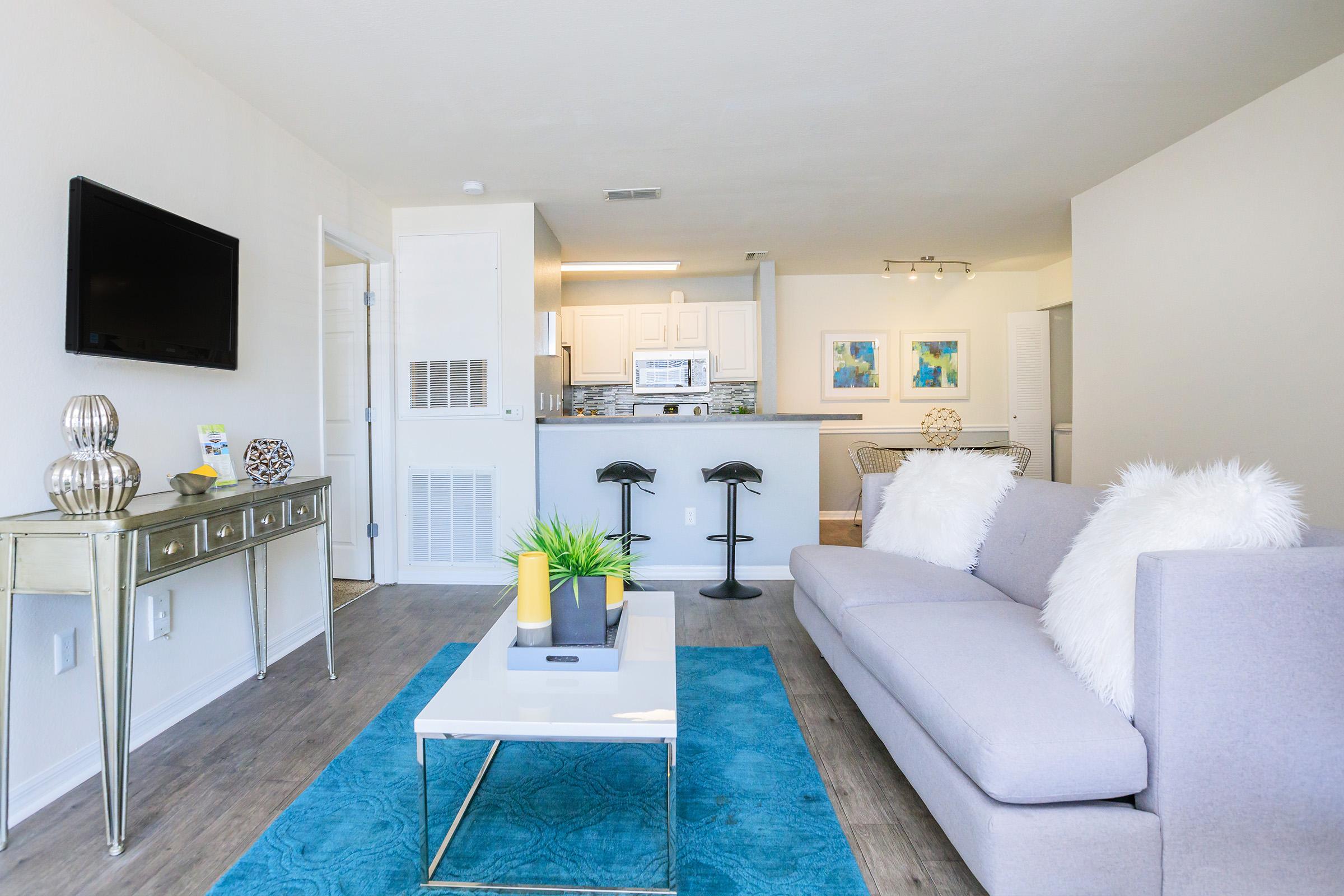 A modern, well-lit living room featuring a gray sofa with fluffy white pillows, a glass coffee table, and a blue area rug. In the background, there’s a kitchen with white cabinetry and stainless steel appliances. A flat-screen TV is mounted on the wall, and two stylish bar stools are positioned at the counter.