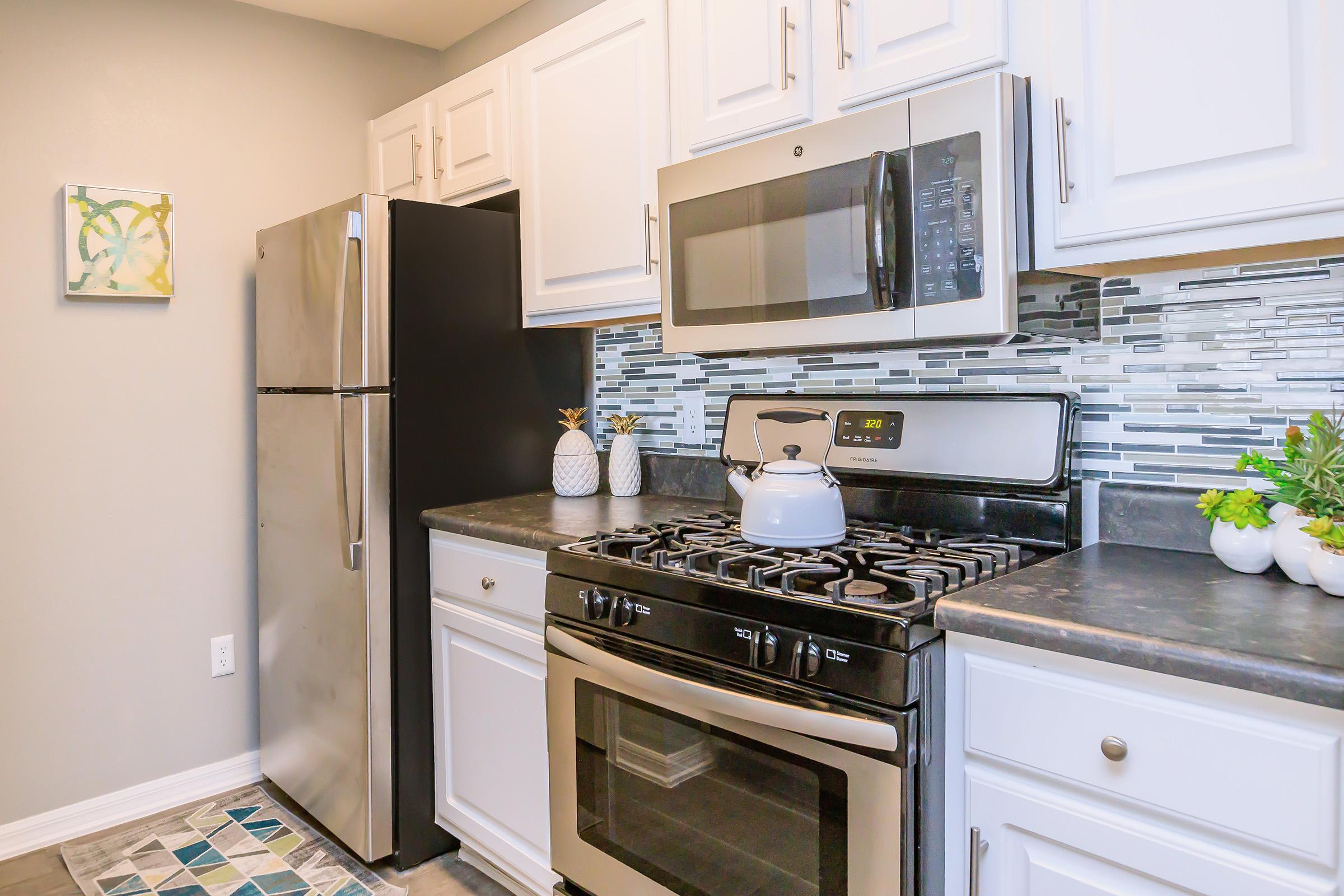 A modern kitchen featuring stainless steel appliances, including a refrigerator and a microwave, alongside a gas stove with an oven. The cabinetry is white with a decorative backsplash in shades of gray and blue. There are small potted plants and a kettle on the stove, adding a touch of warmth to the space.