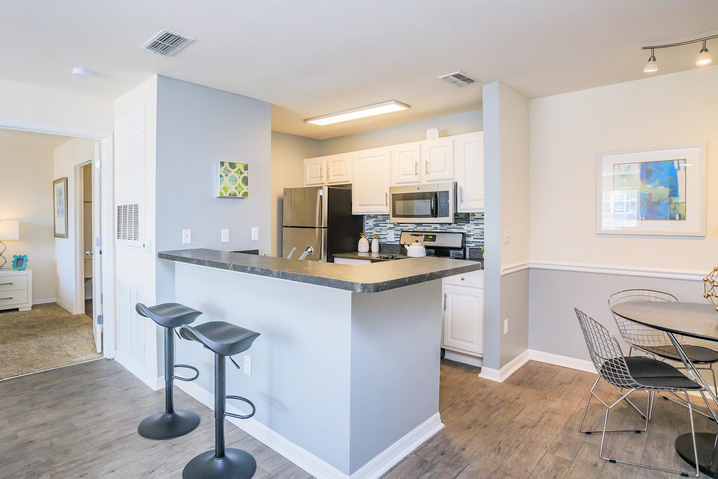 Modern kitchen with white cabinets, stainless steel appliances, and a bar area with two black stools. A dining table with stylish chairs is visible, along with light-colored walls and decorative artwork. The space has an open layout, leading to another room with a cozy ambiance.