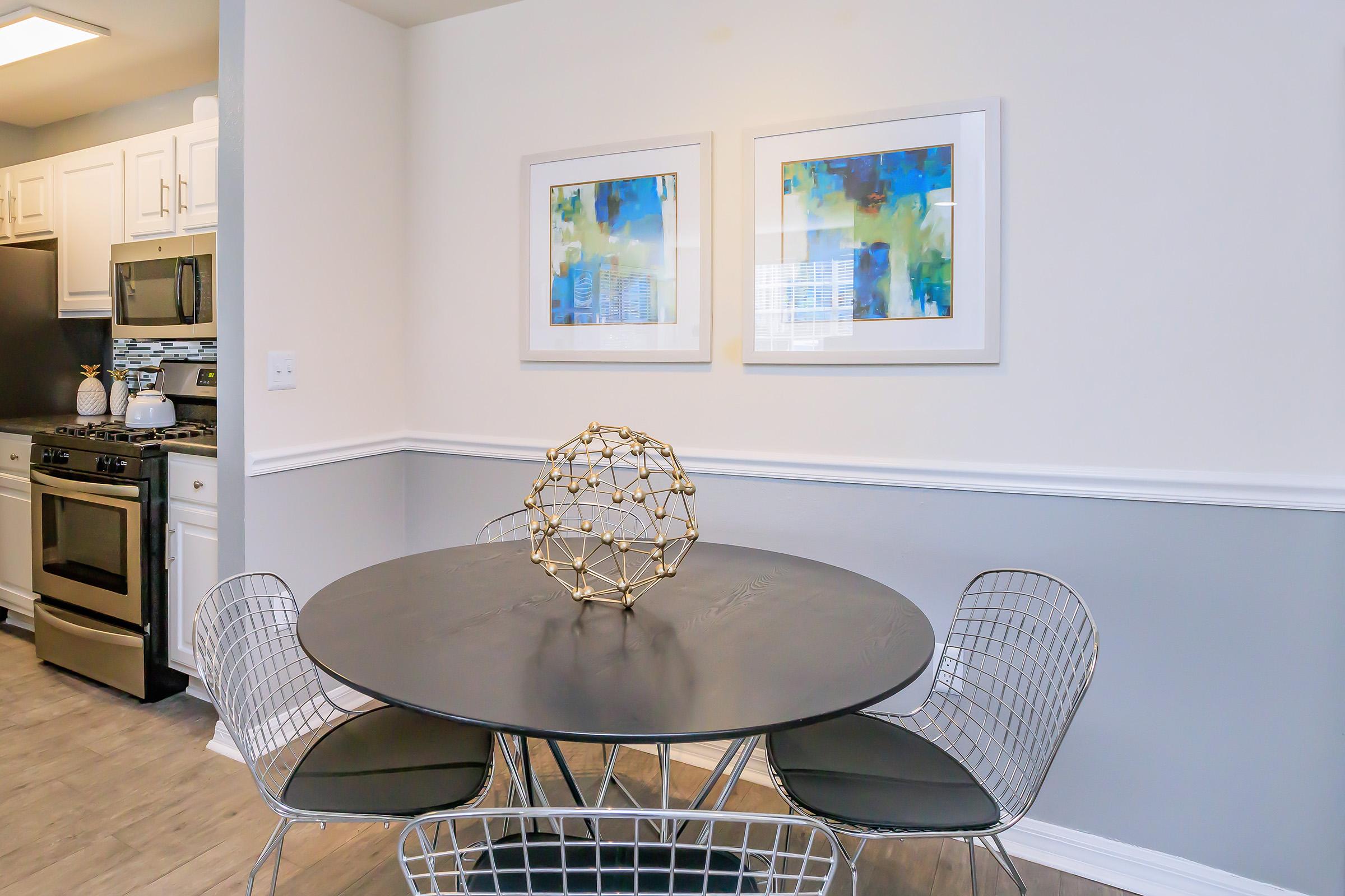 Modern dining area featuring a round black table with wire chairs. On the table, there's a decorative geometric sculpture. The walls are painted in a light color with a darker section, and two framed abstract artworks hang above the table. A kitchen space is visible in the background with stainless steel appliances.