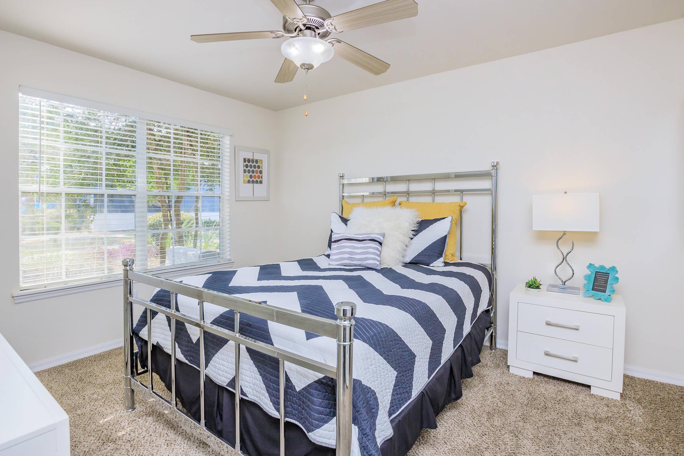 A cozy bedroom featuring a metal bed frame with a blue and white chevron-patterned comforter. The room is brightened by a window with blinds and includes a bedside table with a lamp and decorative items. A ceiling fan hangs above, adding a modern touch to the inviting space.