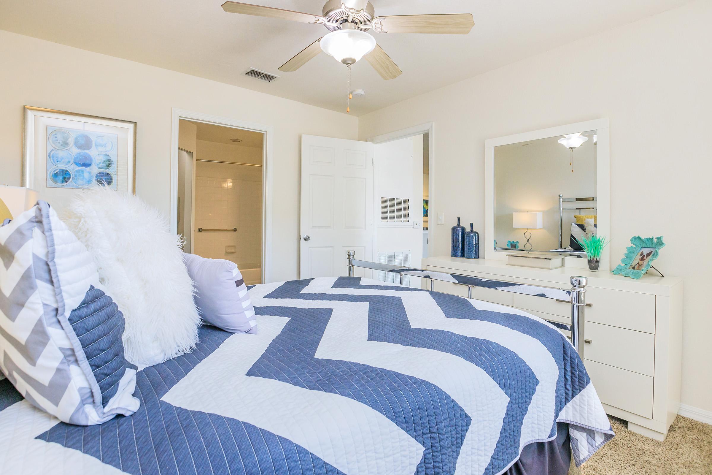 A cozy bedroom featuring a bed with a chevron-patterned comforter in navy blue and white. A ceiling fan hangs above, and there's a dresser with decorative items. The walls are painted a soft neutral color, and a mirror reflects the light. A bathroom door is visible in the background.