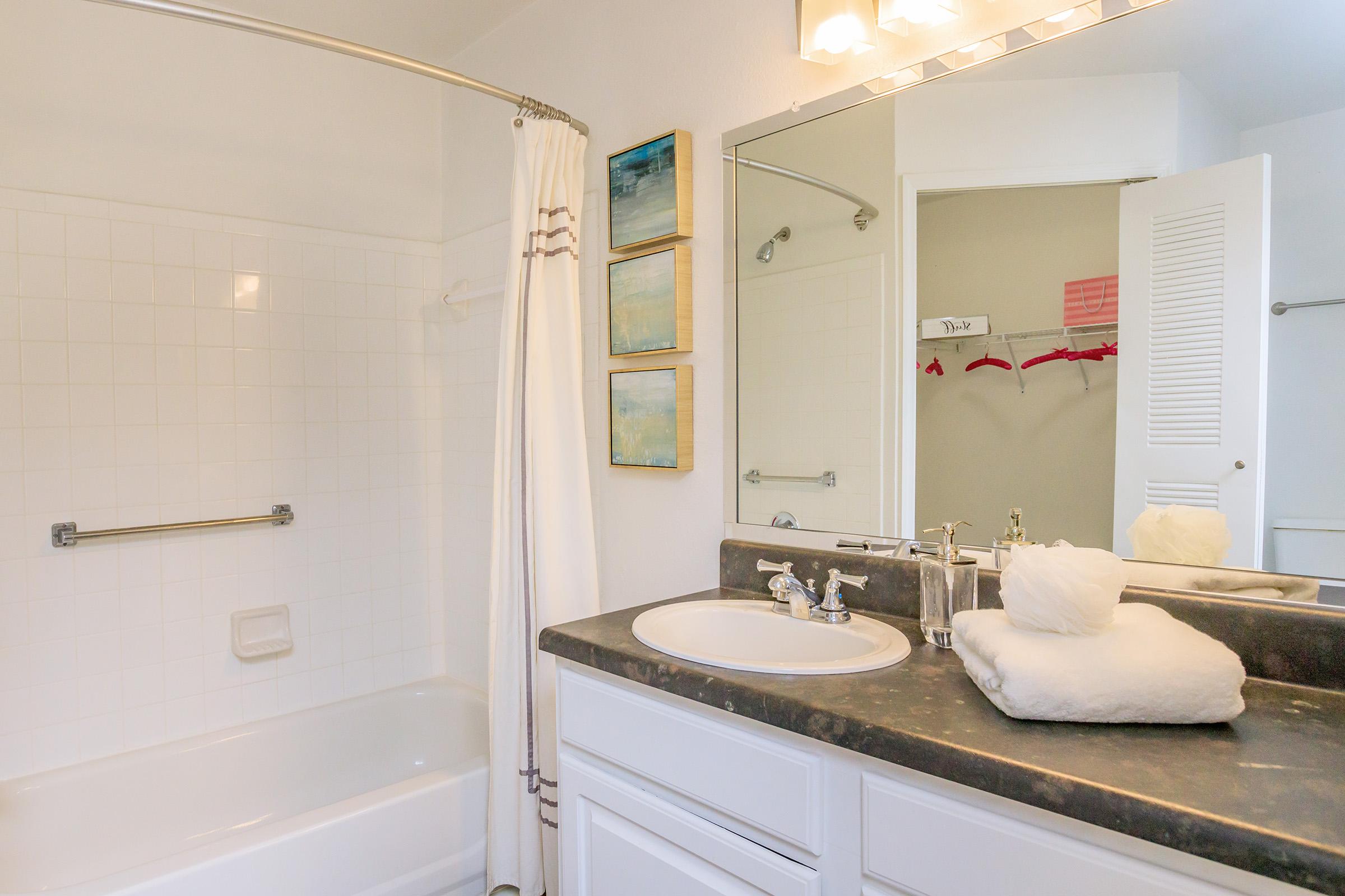 A clean and modern bathroom featuring a bathtub with a shower curtain, a sink with toiletries, and neatly arranged towels. Wall decor includes two abstract paintings. A lighted mirror reflects the bright, spacious design, and a closet door is visible in the background.