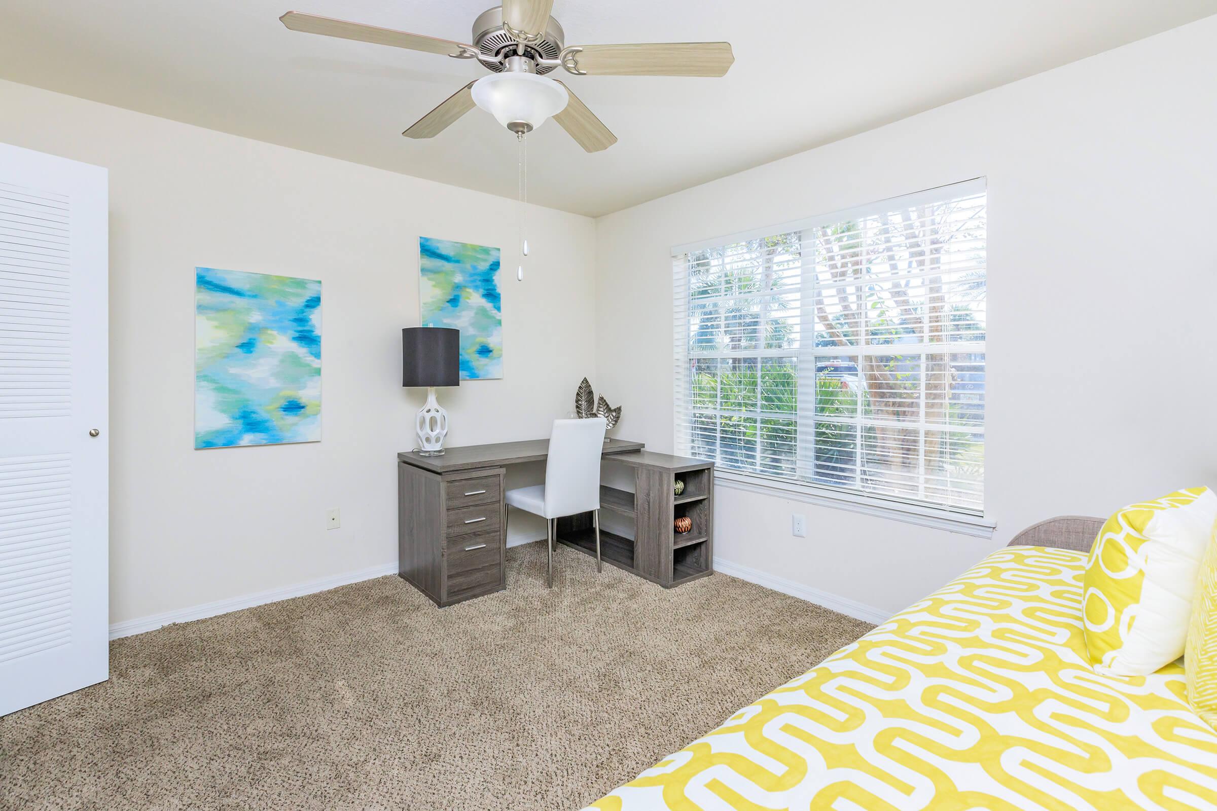 A bright and airy bedroom featuring a ceiling fan, a large window with natural light, a desk with a lamp, and two abstract wall art pieces in blue and green. The room has light-colored walls and carpet, with a cozy yellow and white patterned throw on the bed.