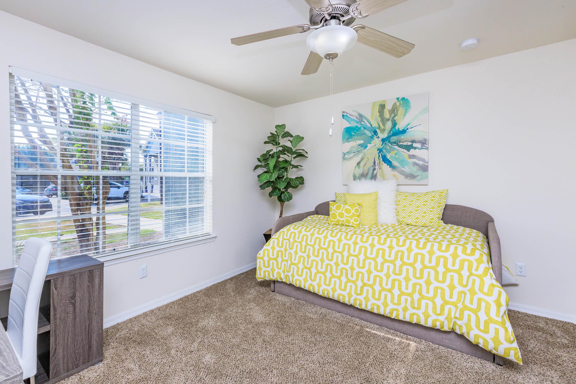 A bright and cozy bedroom featuring a daybed with yellow and white patterned bedding, a decorative pillow, and a fluffy white throw. There is a small desk and a window with white blinds allowing natural light to flood the room. A green plant adds a touch of nature to the space.